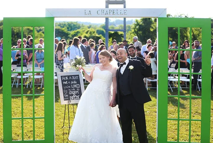 A newlywed couple stands under a green archway labeled 'La Chapelle' at their outdoor wedding, holding hands and smiling, with guests seated behind them.