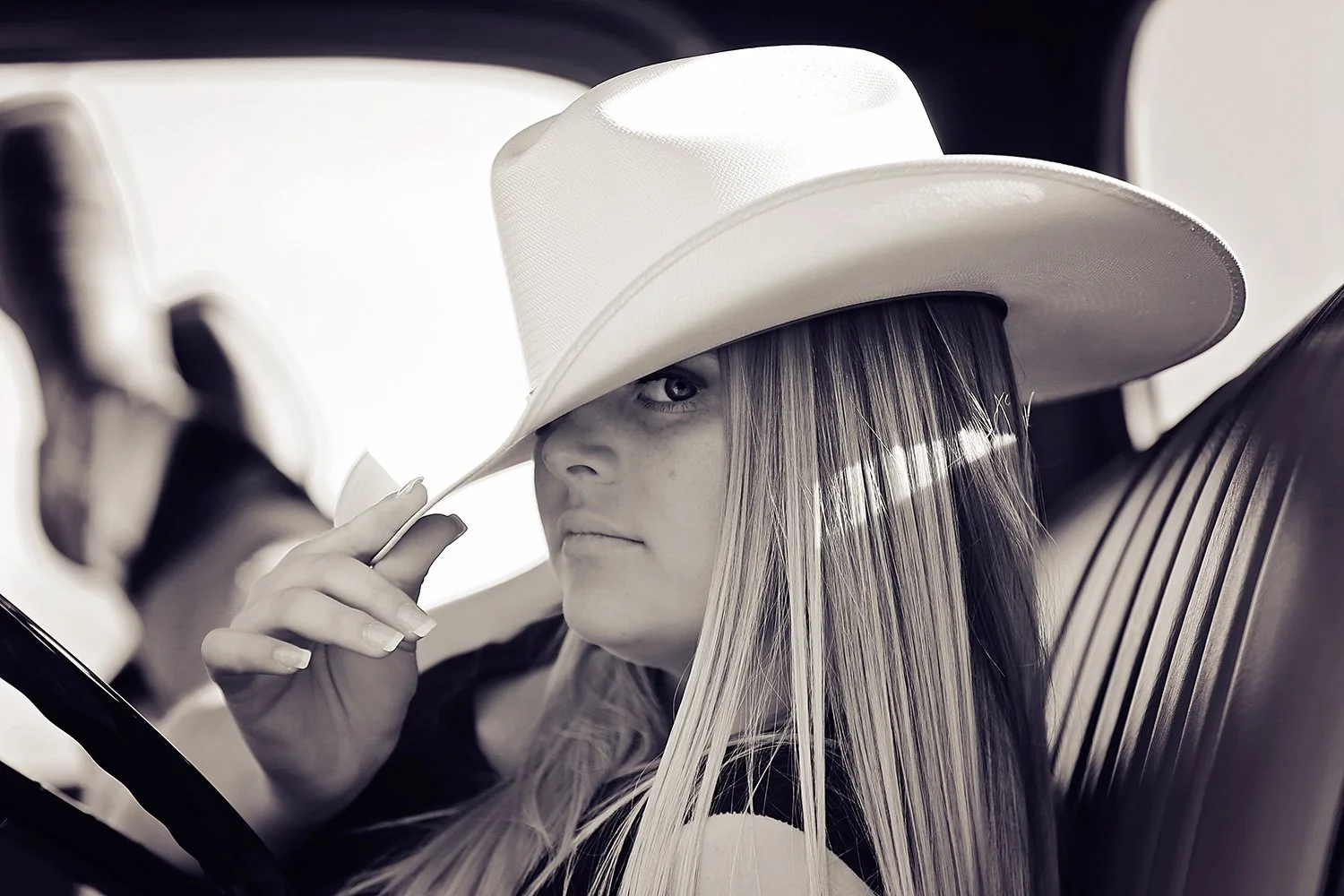 Black and white photo of a woman wearing a large cowboy hat and sitting in a vehicle. She is slightly tilting her hat and looking at the camera with one eye visible, while her long hair falls over her shoulder.