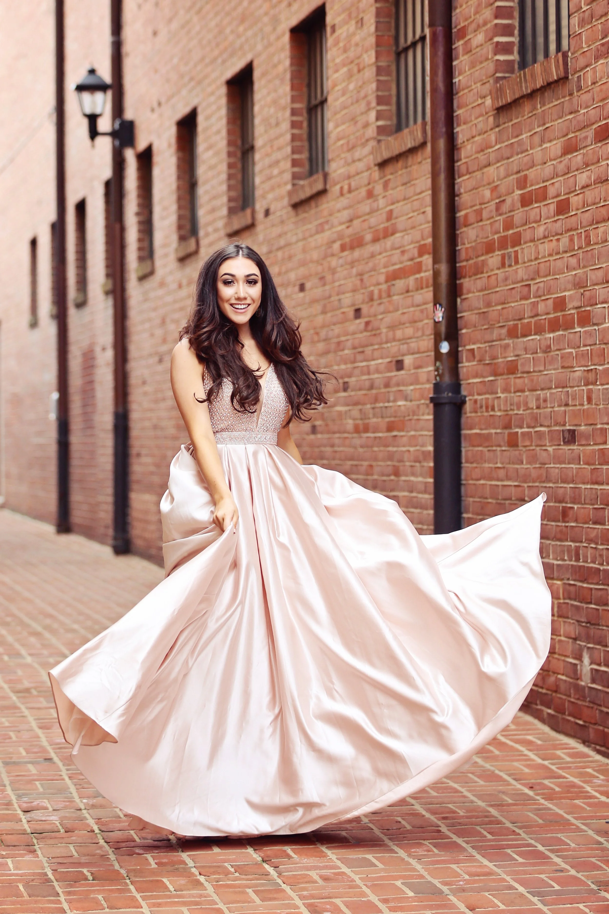 A young woman with long dark hair wearing a pink satin ball gown, twirling on a brick sidewalk next to a brick building with windows and vintage-style street lamps.