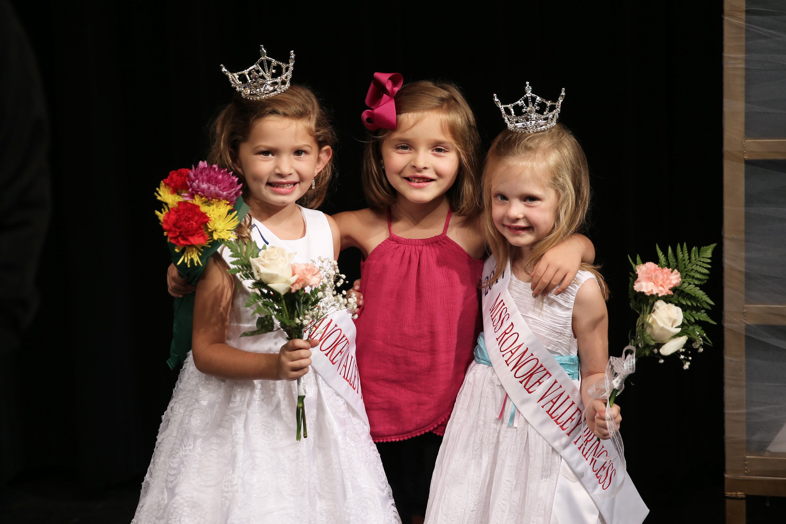 Three young girls at a pageant, two wearing crowns and sashes, holding bouquets of flowers, smiling for a photo, with a dark background.