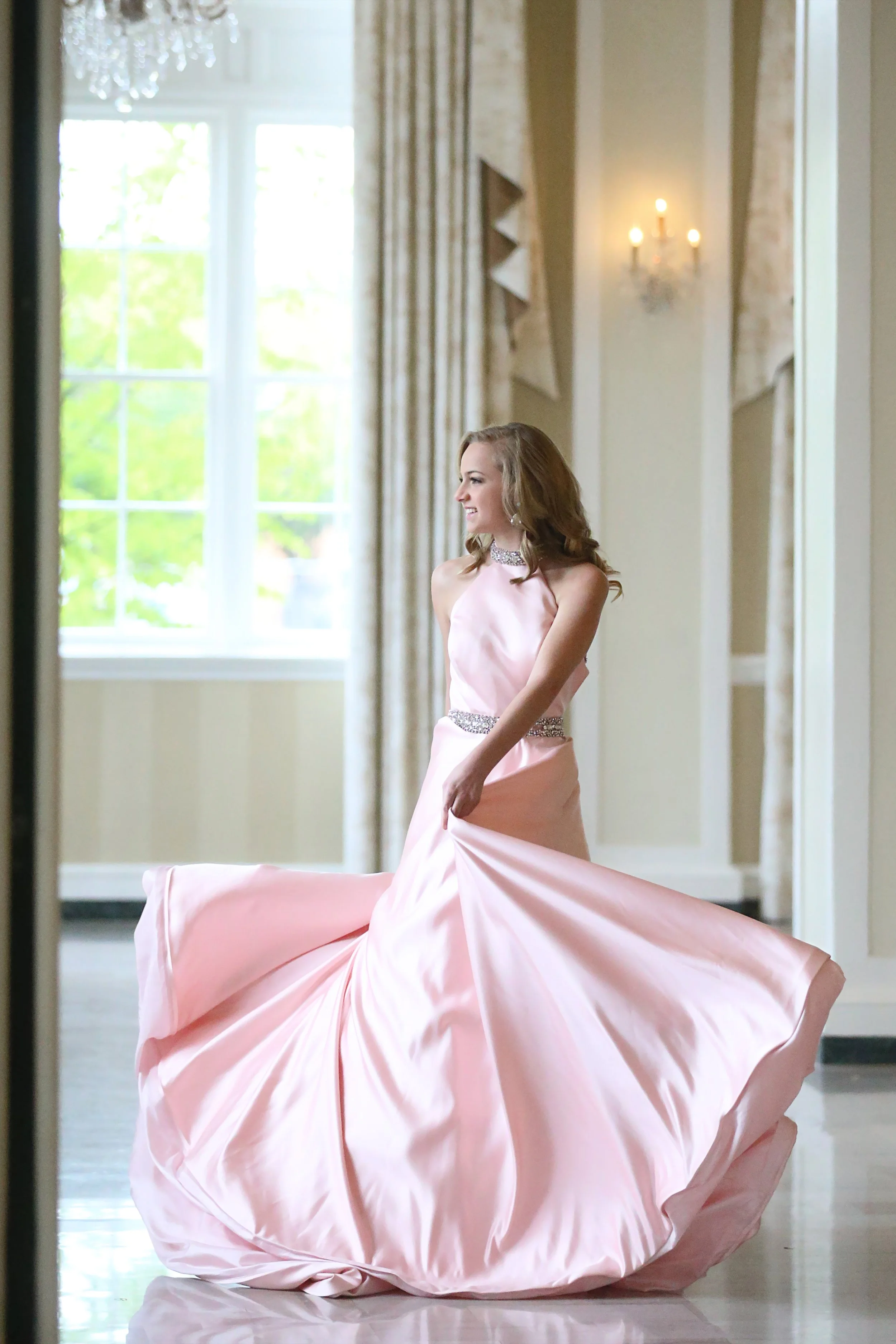 A woman in a pink satin gown, smiling and twirling inside a well-lit room with large windows and elegant decor.