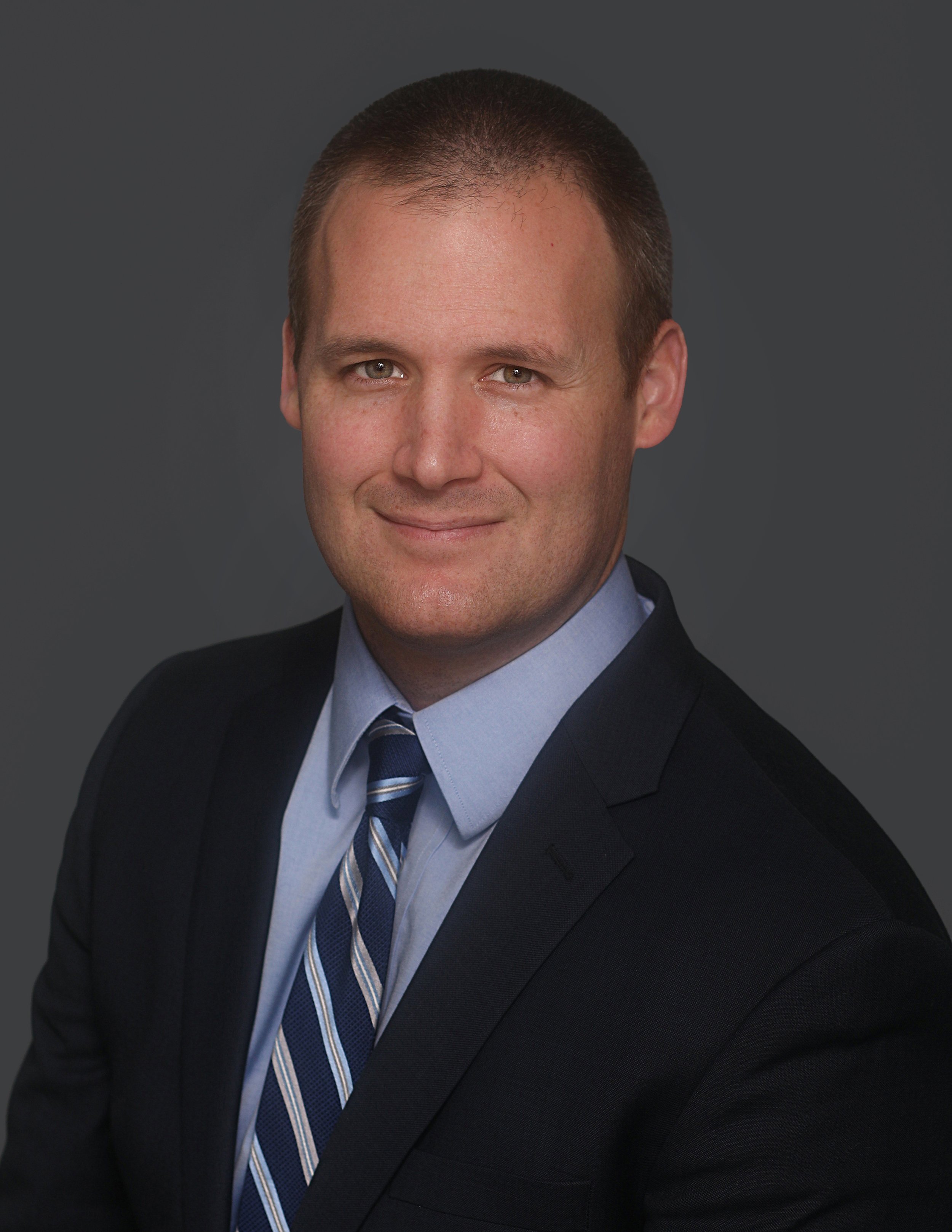 Professional headshot of a man in a dark suit, light blue shirt, and striped tie, against a dark gray background.