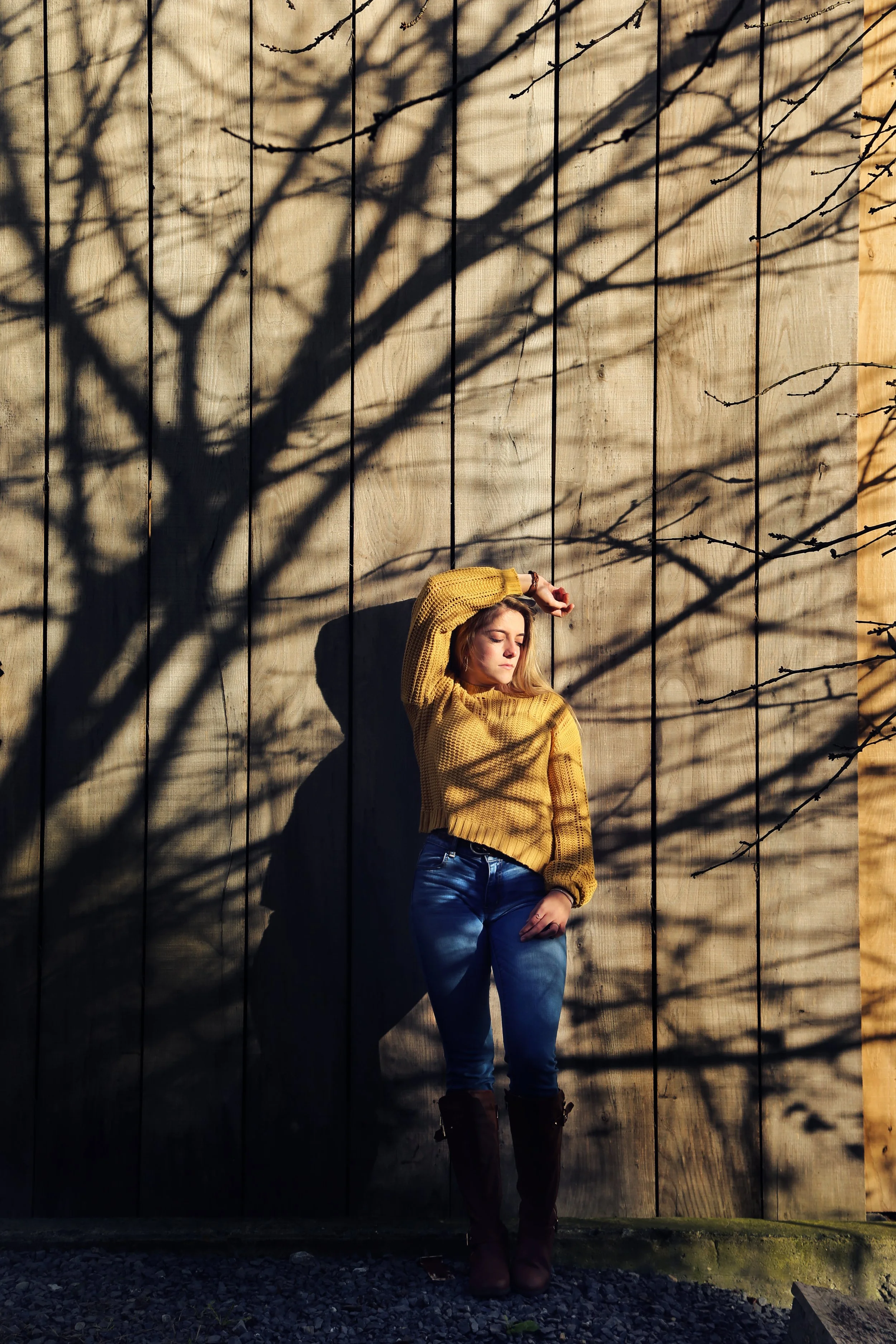 A woman standing outdoors against a wooden wall with shadows of tree branches cast on it, wearing a yellow sweater and jeans.