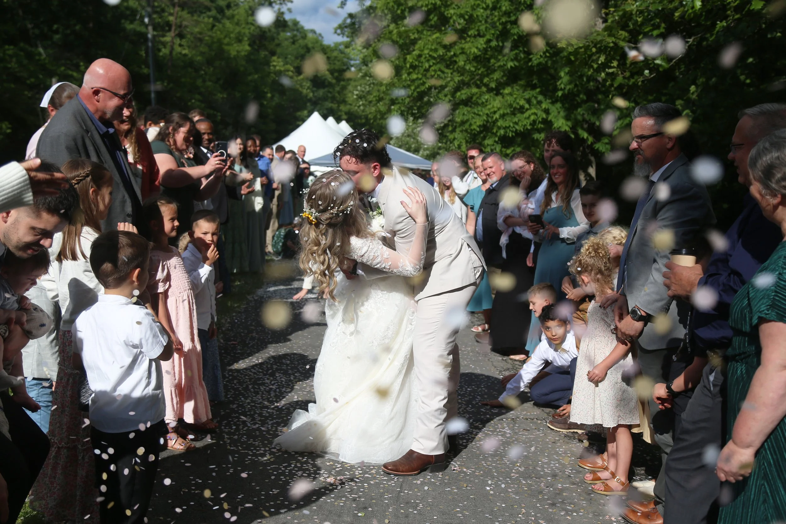 A wedding scene with a bride and groom kissing on a pathway, surrounded by a crowd of guests tossing confetti, outdoors with green trees and white tents in the background.