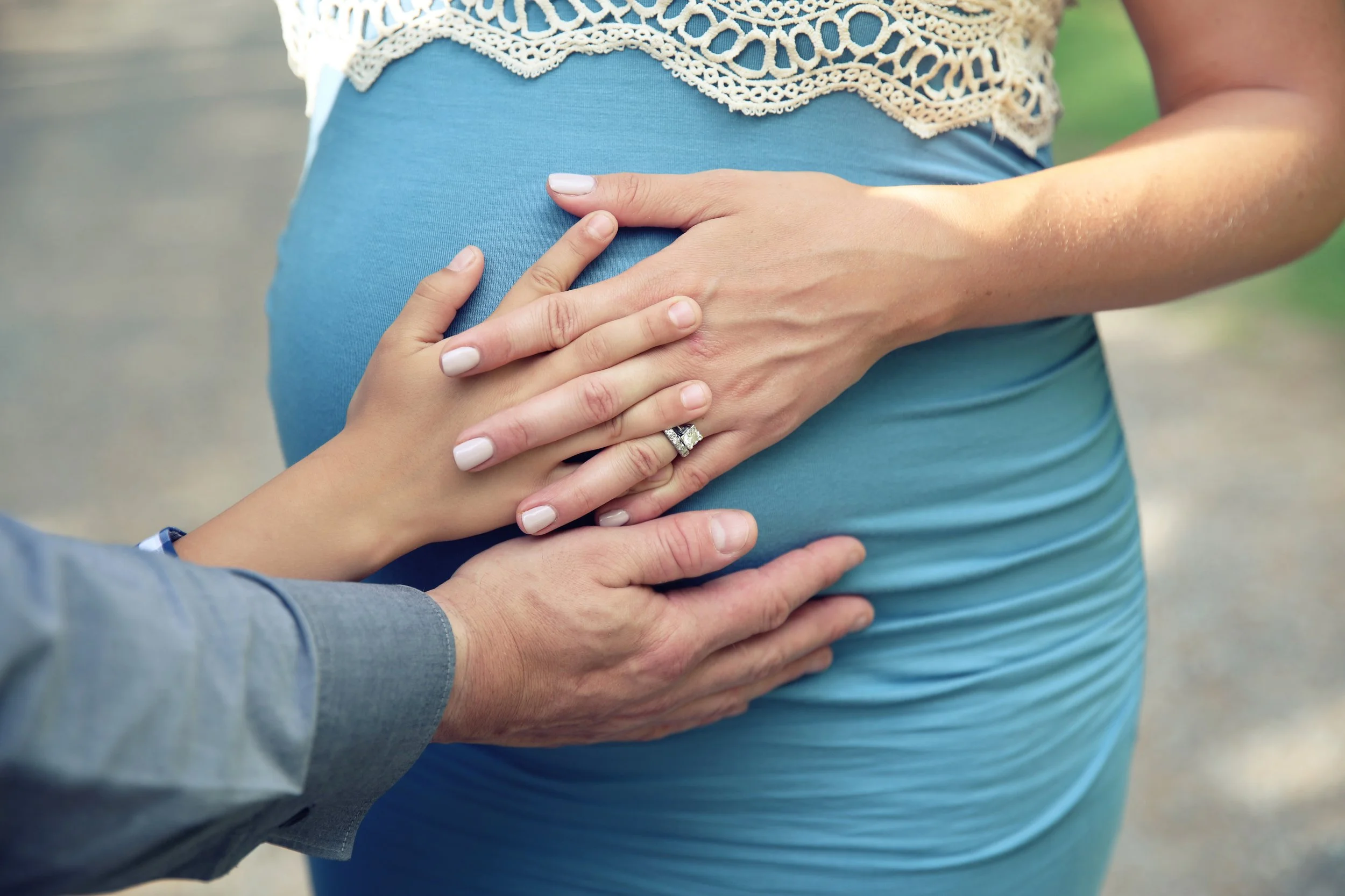 Close-up of a pregnant woman's belly with a man and woman’s hands gently resting on her stomach. The woman has manicured nails and is wearing an engagement ring, and the man is dressed in a gray shirt. The photo captures an intimate, caring moment outdoors.