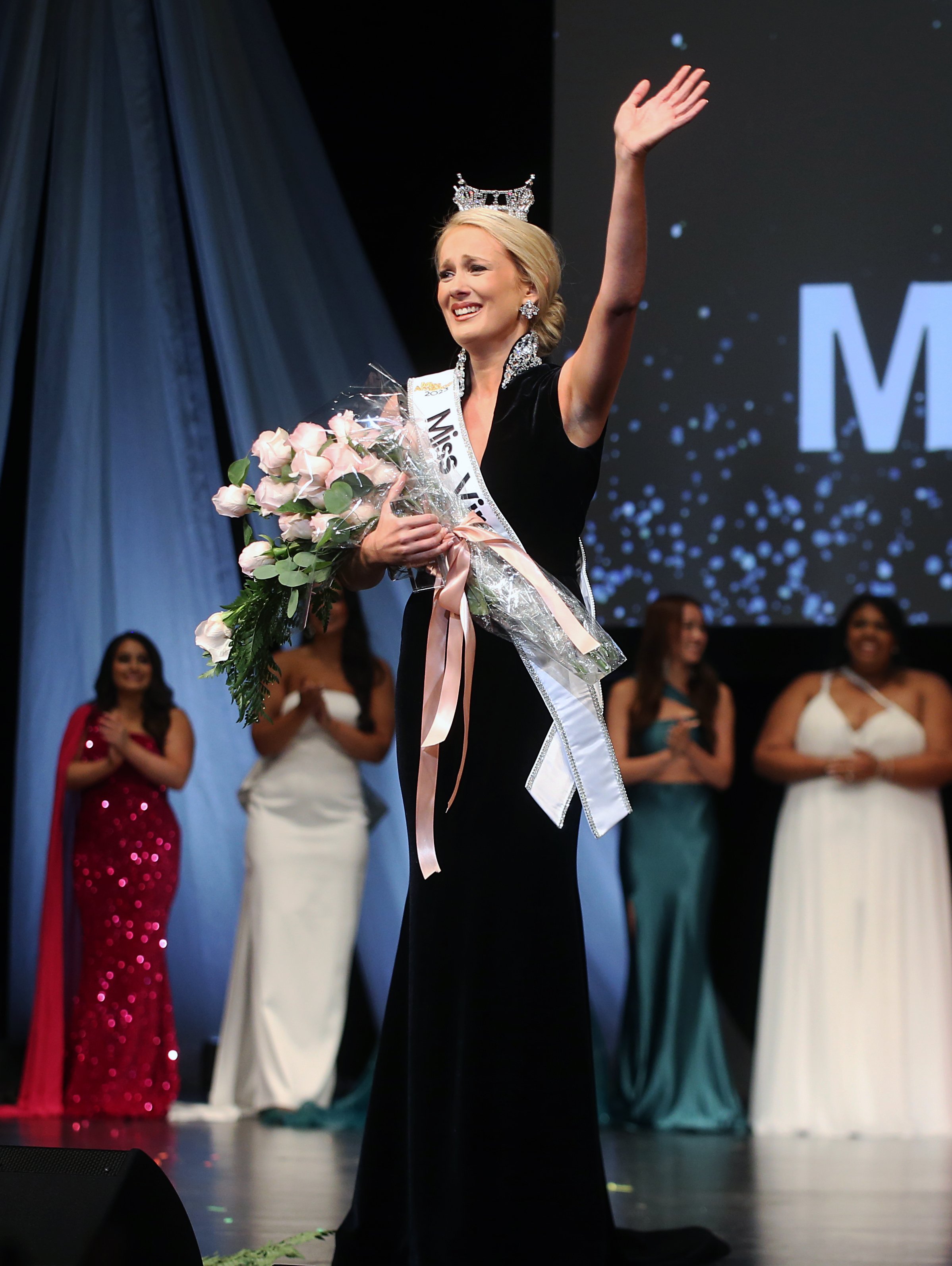 Miss USA winner wearing a crown and sash, holding a bouquet of pink roses, waving on stage with three women in evening gowns behind her.