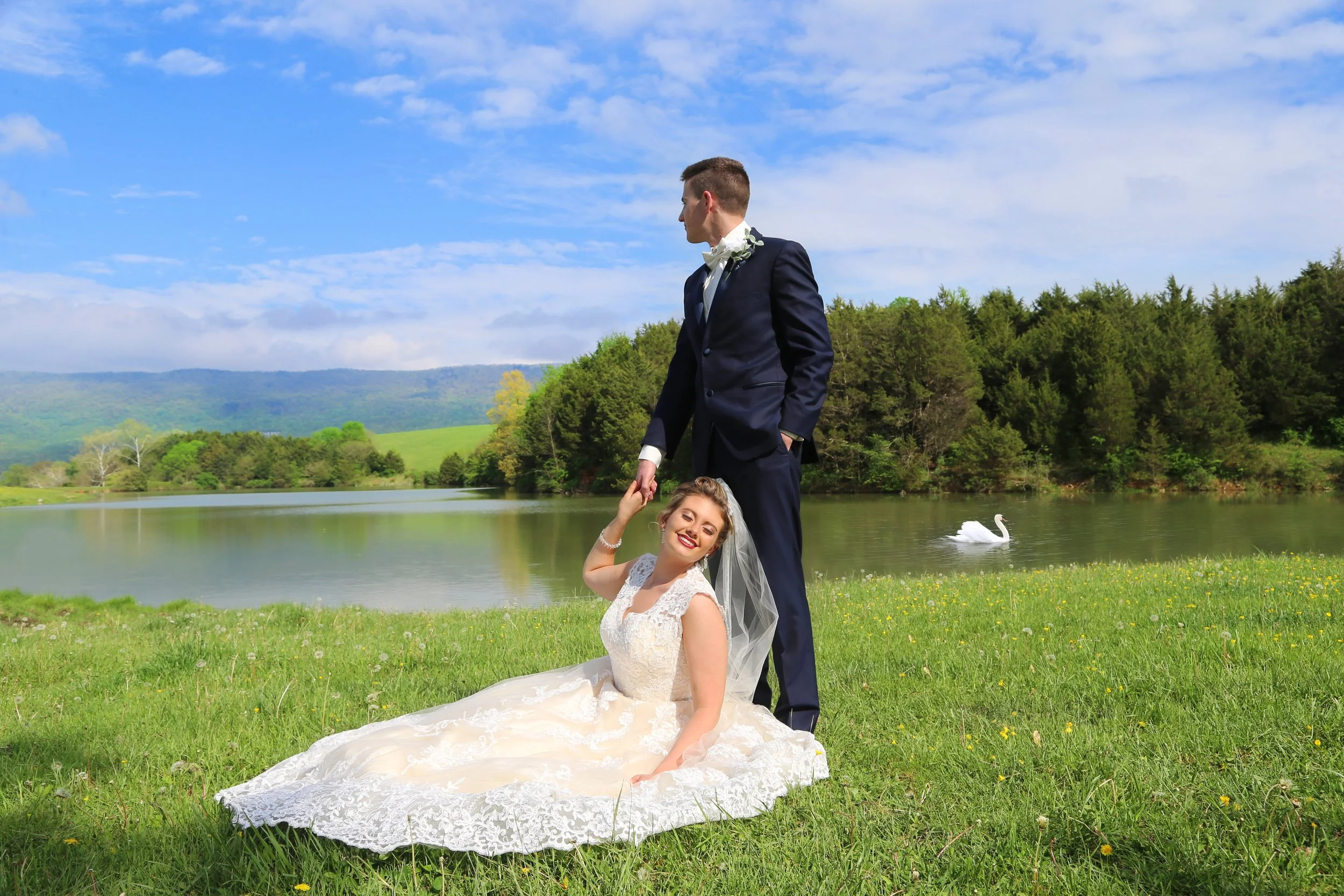 Bride sitting on grass next to groom standing near a lake with swan, trees, and mountains in the background on a sunny day.