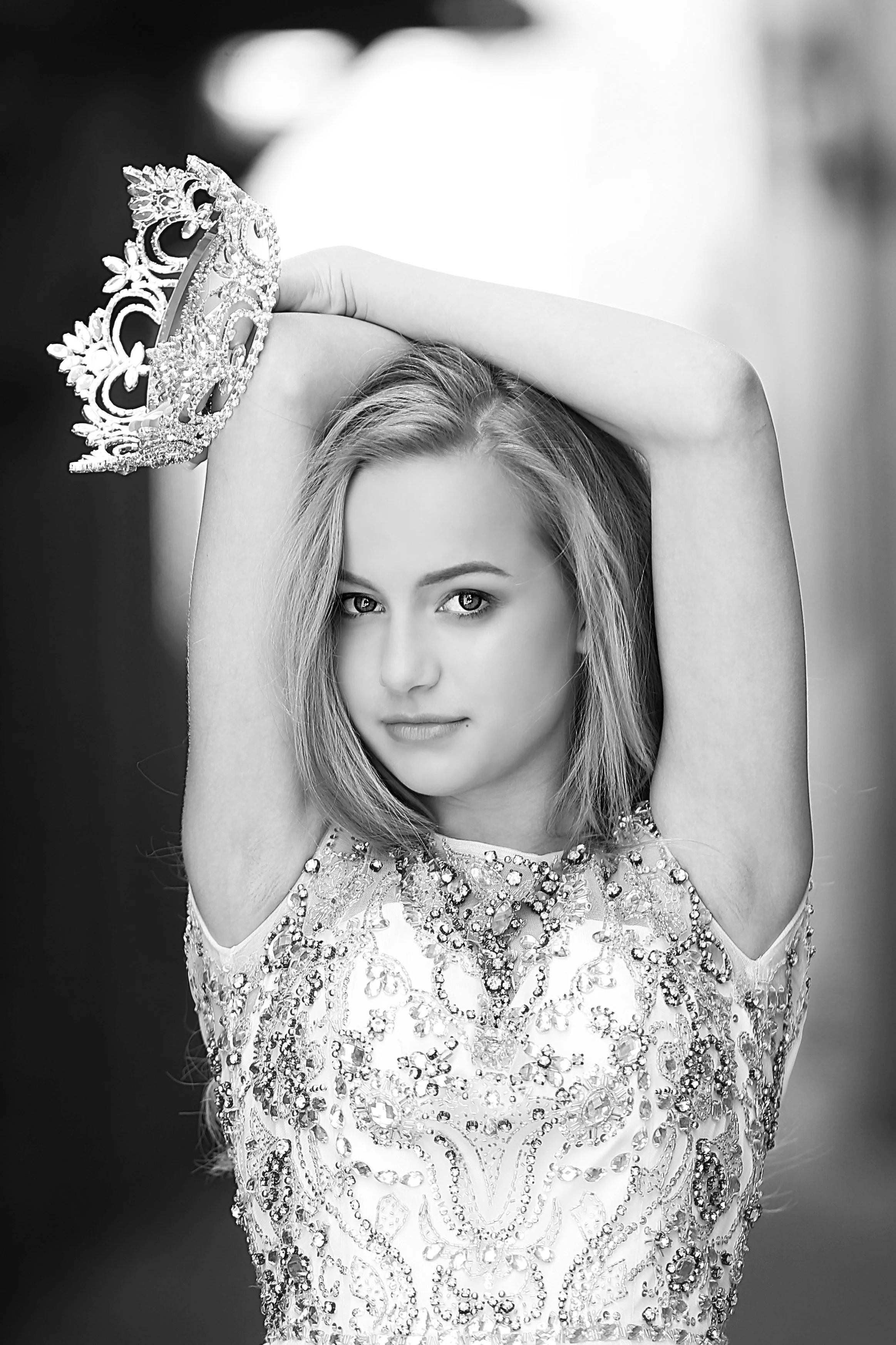 A young woman with shoulder-length hair, wearing a beaded or sequined dress, holding a tiara above her head with her right hand, in a black-and-white photo.