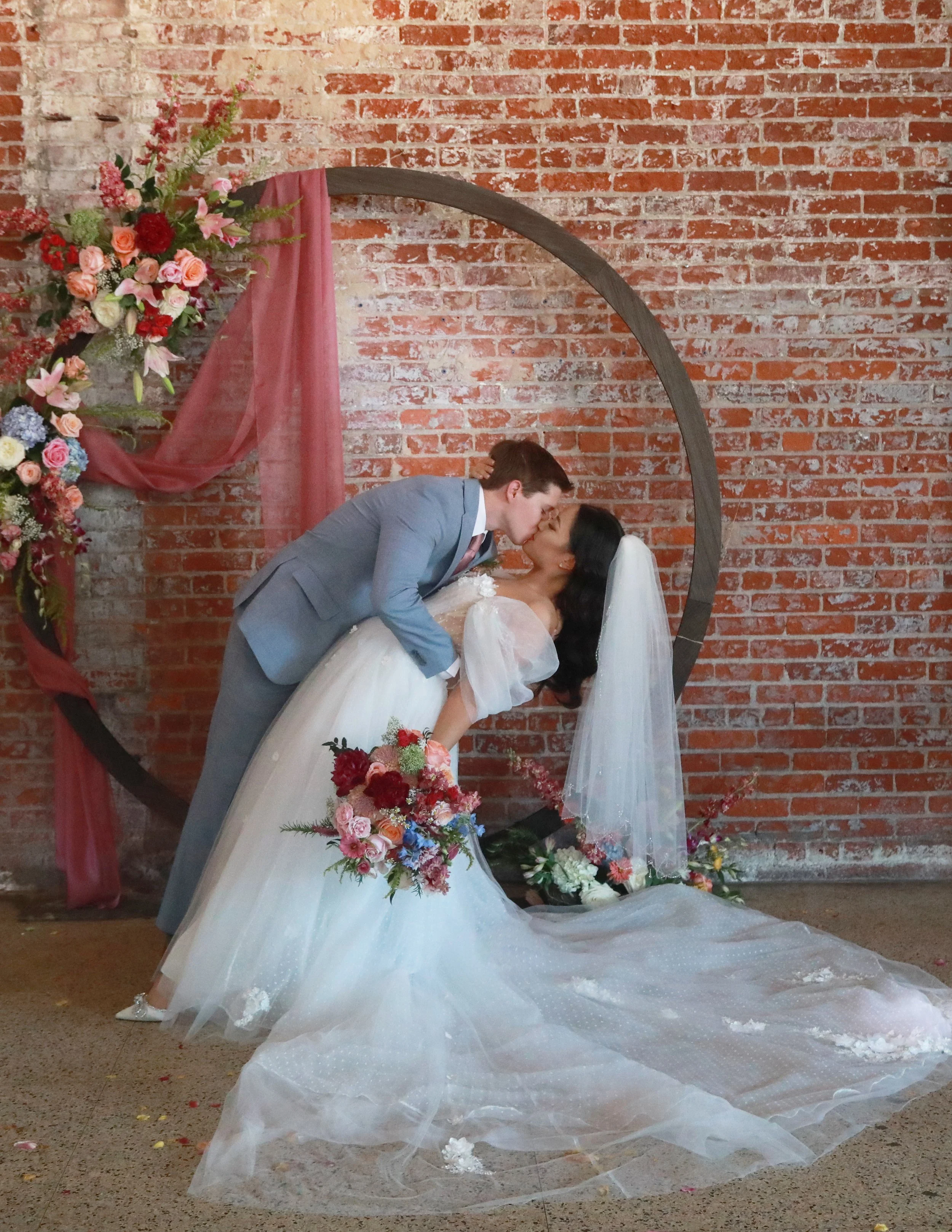 A bride and groom sharing a kiss during their wedding, with the groom dipping the bride, who is holding a bouquet of flowers, in front of a brick wall and floral decorations.