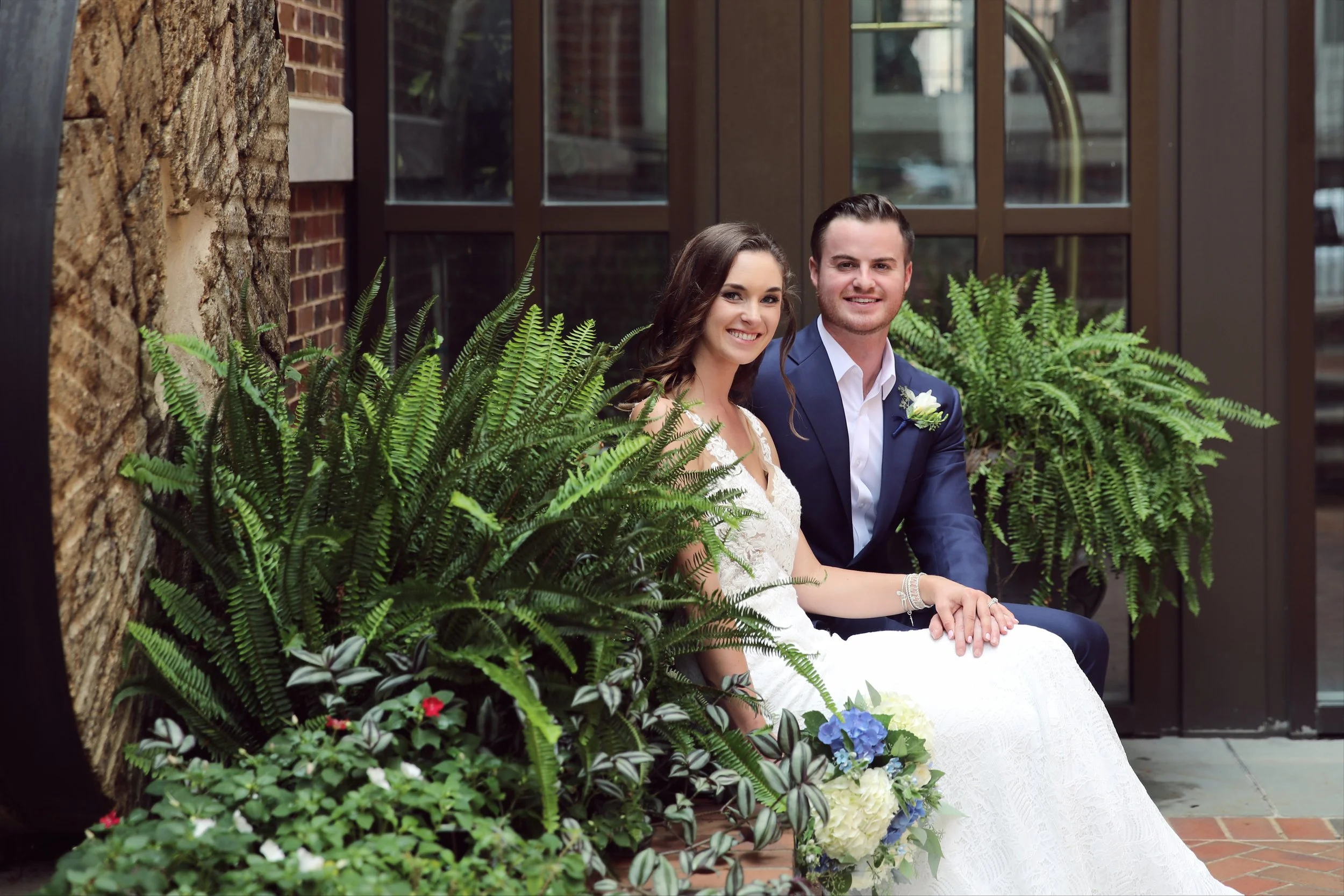 A bride and groom sitting on a bench outside, surrounded by green plants and flowers, smiling at the camera.