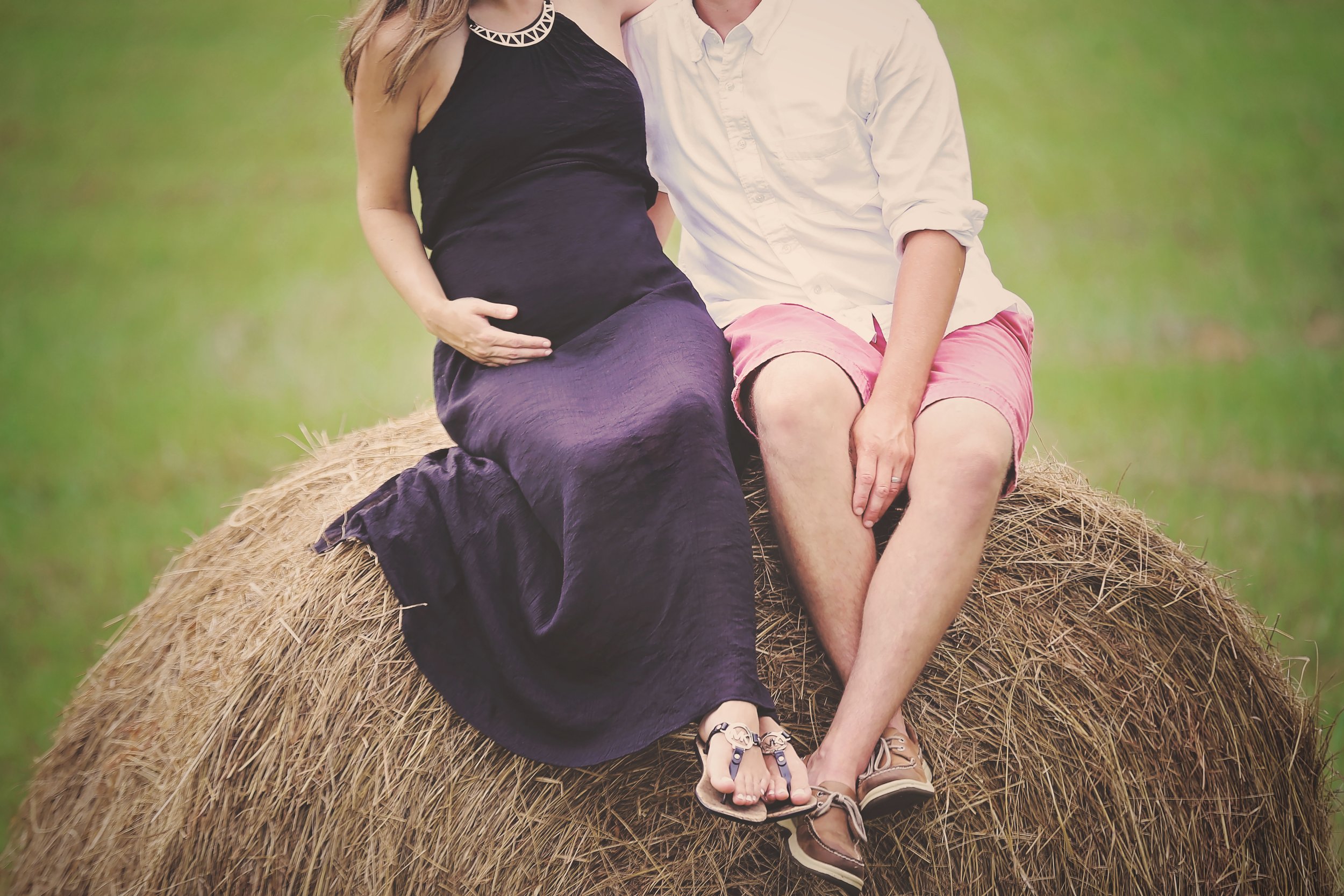 A pregnant woman and a man sitting on a hay bale outdoors, holding hands and displaying her baby bump.