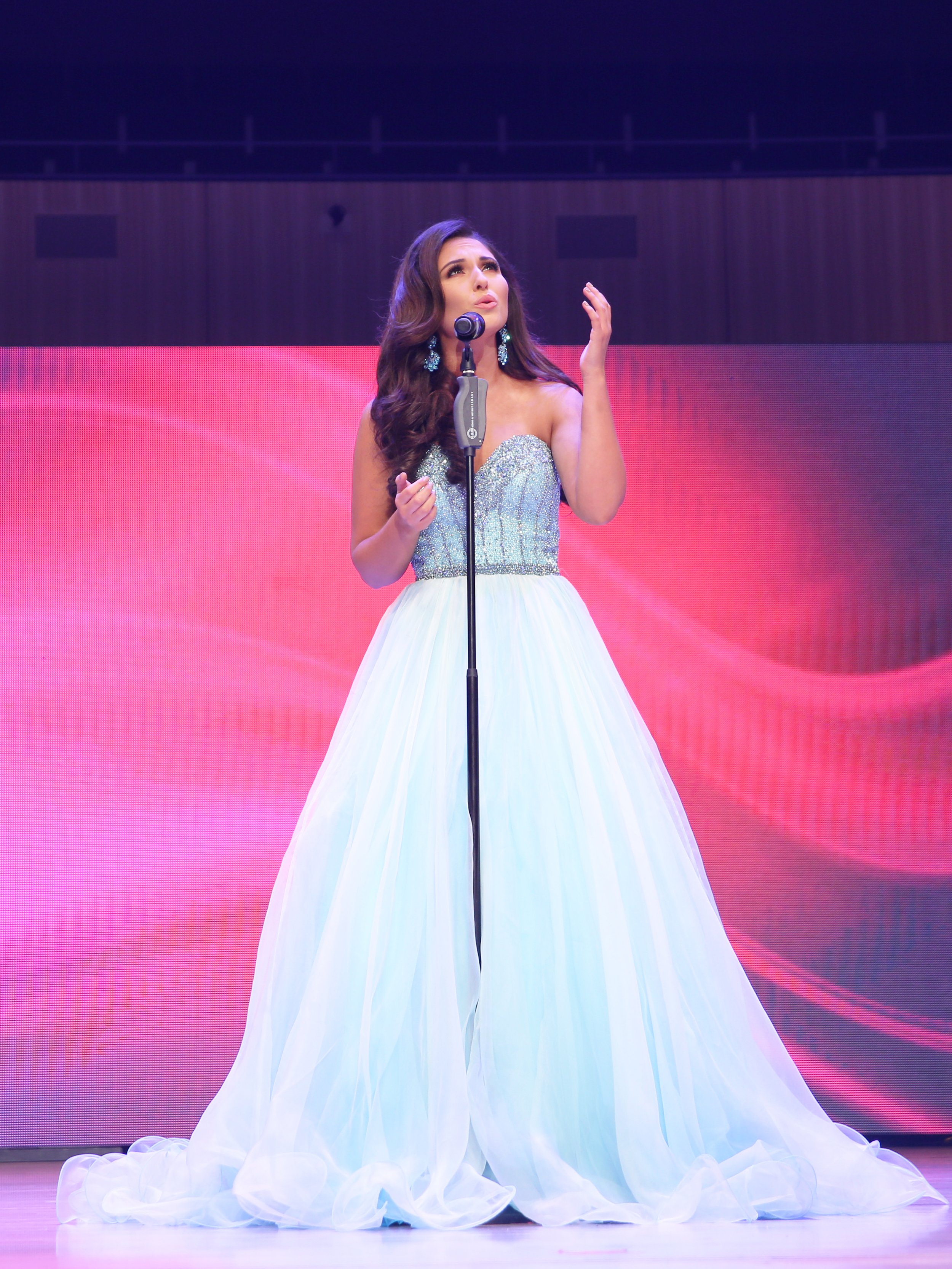A young woman in a strapless, sparkling, silver and white gown singing into a microphone on stage with pink and purple background lighting.