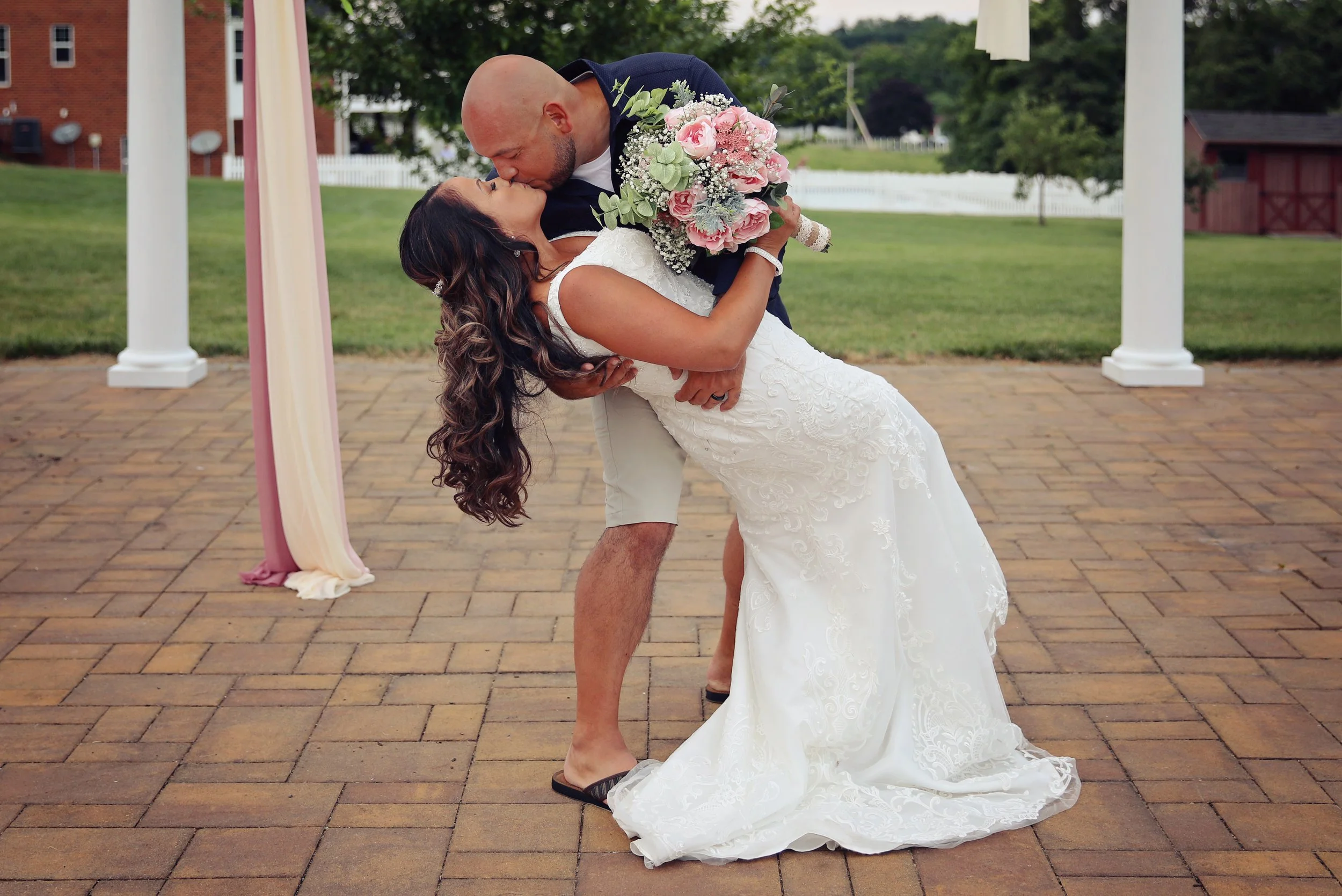 A bride and groom share a kiss during their outdoor wedding ceremony. The groom is wearing shorts and a dark shirt, and the bride is in a white lace wedding gown holding a bouquet of pink and white flowers. They are standing on a brick patio with a decorated arch in the background.