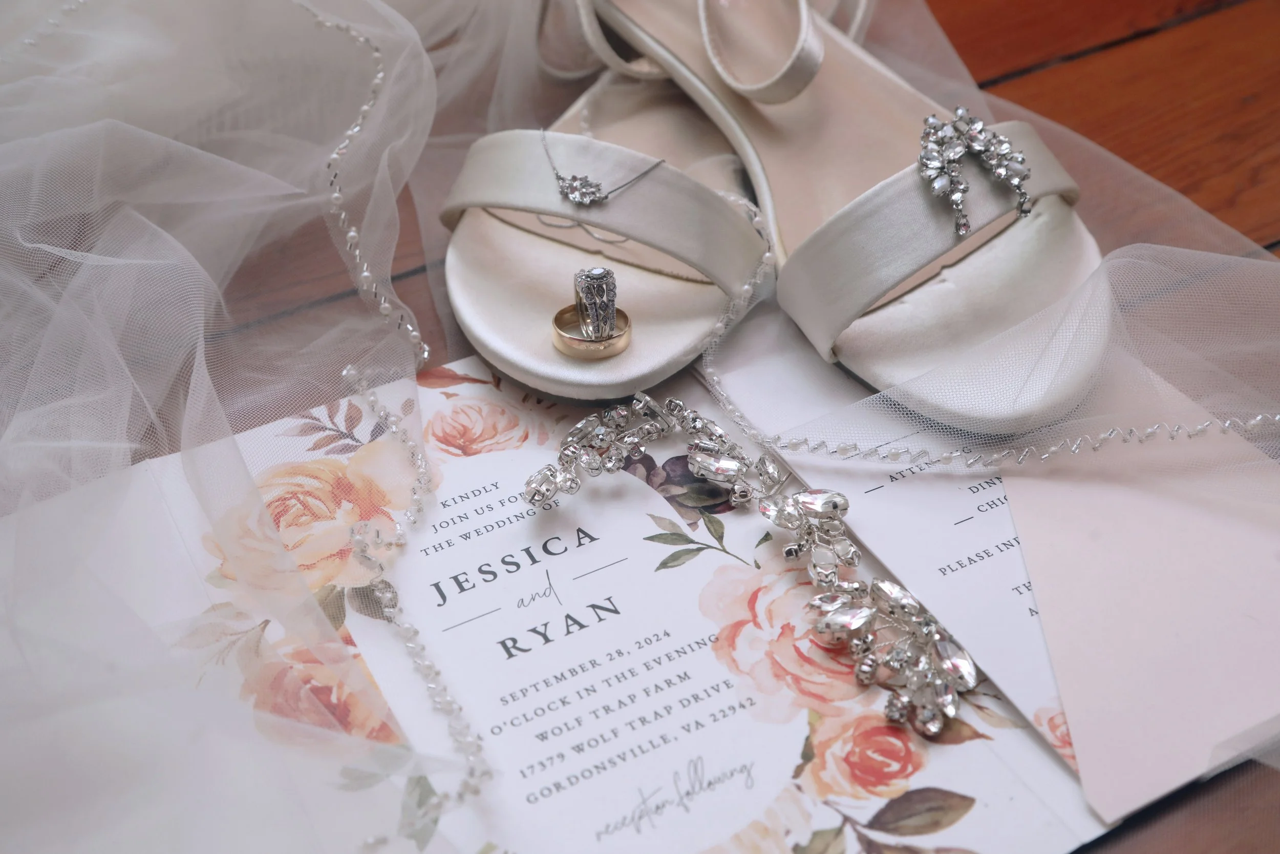 Wedding invitation with jewelry including a ring, necklace, bracelet, and earrings, along with white satin shoes and a veil on a wooden surface.