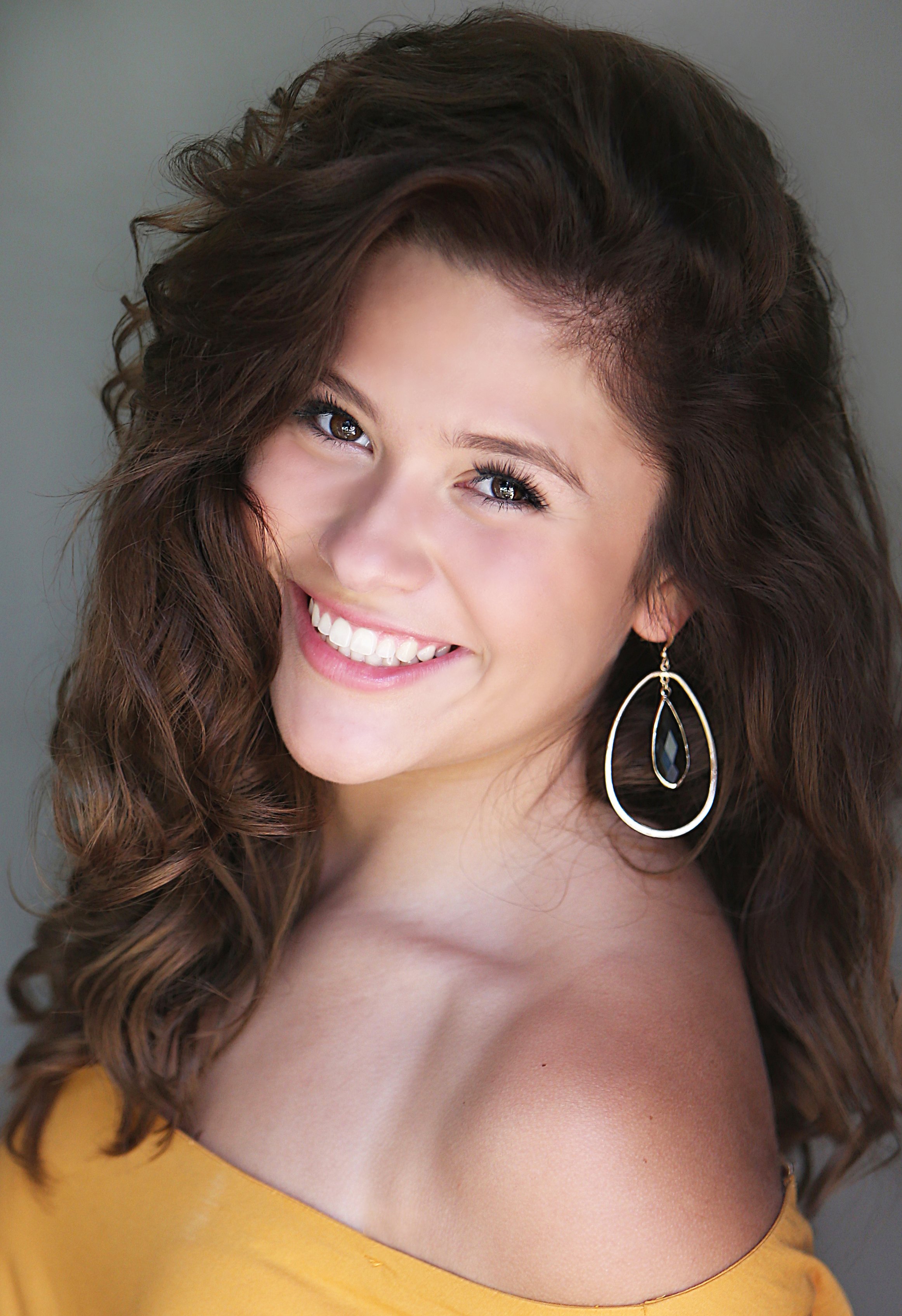 A young woman with long, curly brown hair, smiling, wearing a yellow top and large teardrop-shaped earrings with black accents.