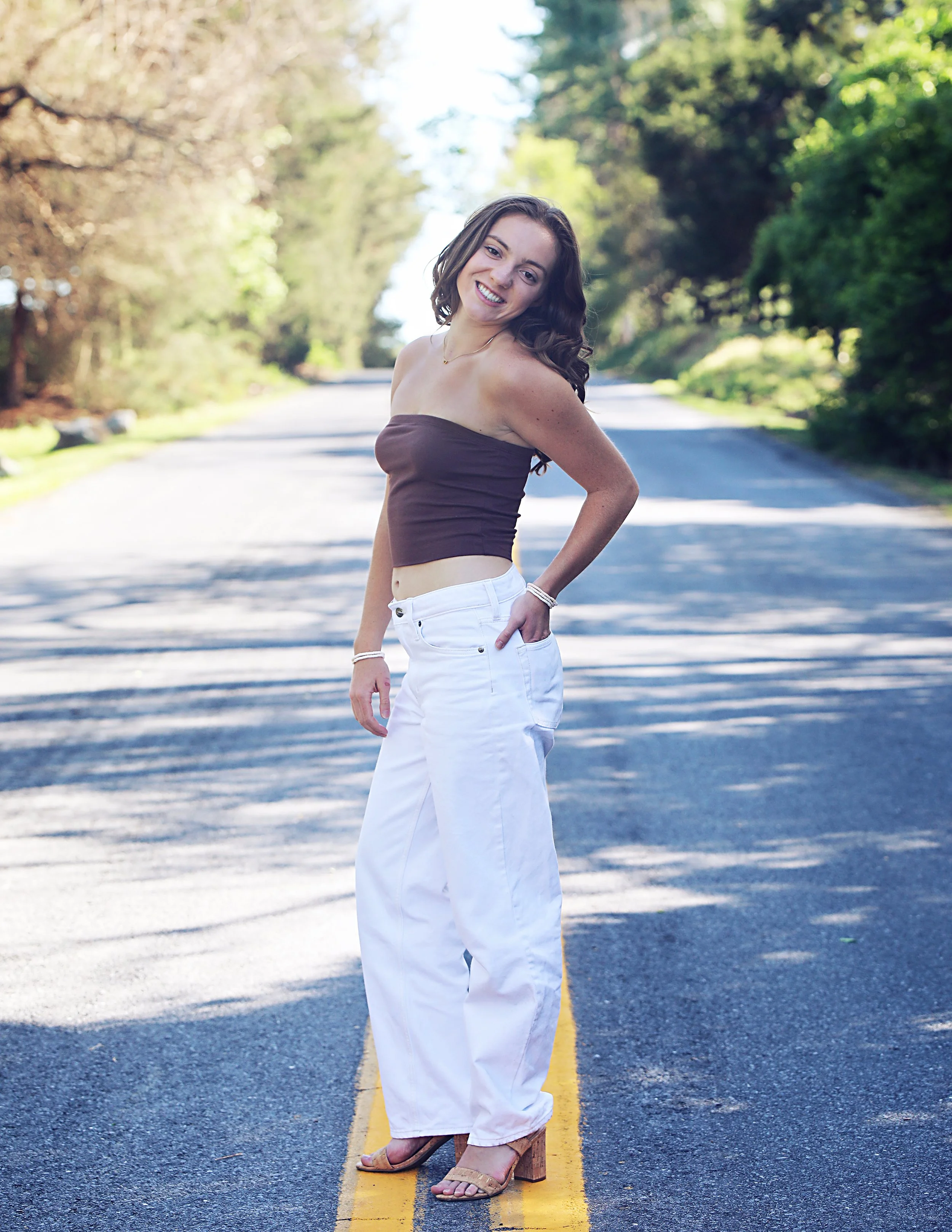 A woman standing on an empty city street with trees in the background, smiling and posing for the photo, wearing a brown strapless top and white pants.
