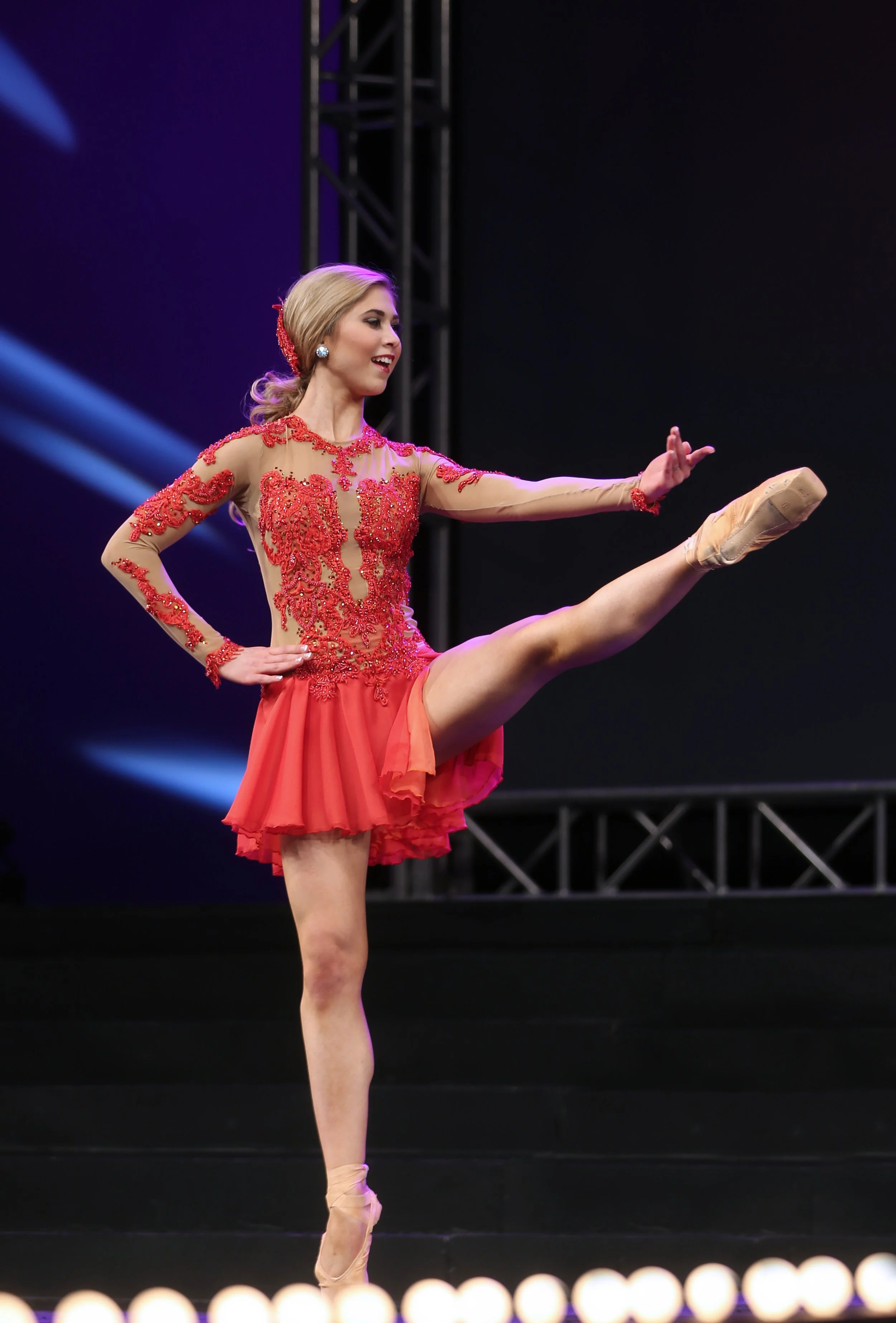 A female ballet dancer performing on stage, wearing a red and beige costume with embroidery and a short skirt, executing an extended leg pose on pointe shoes.