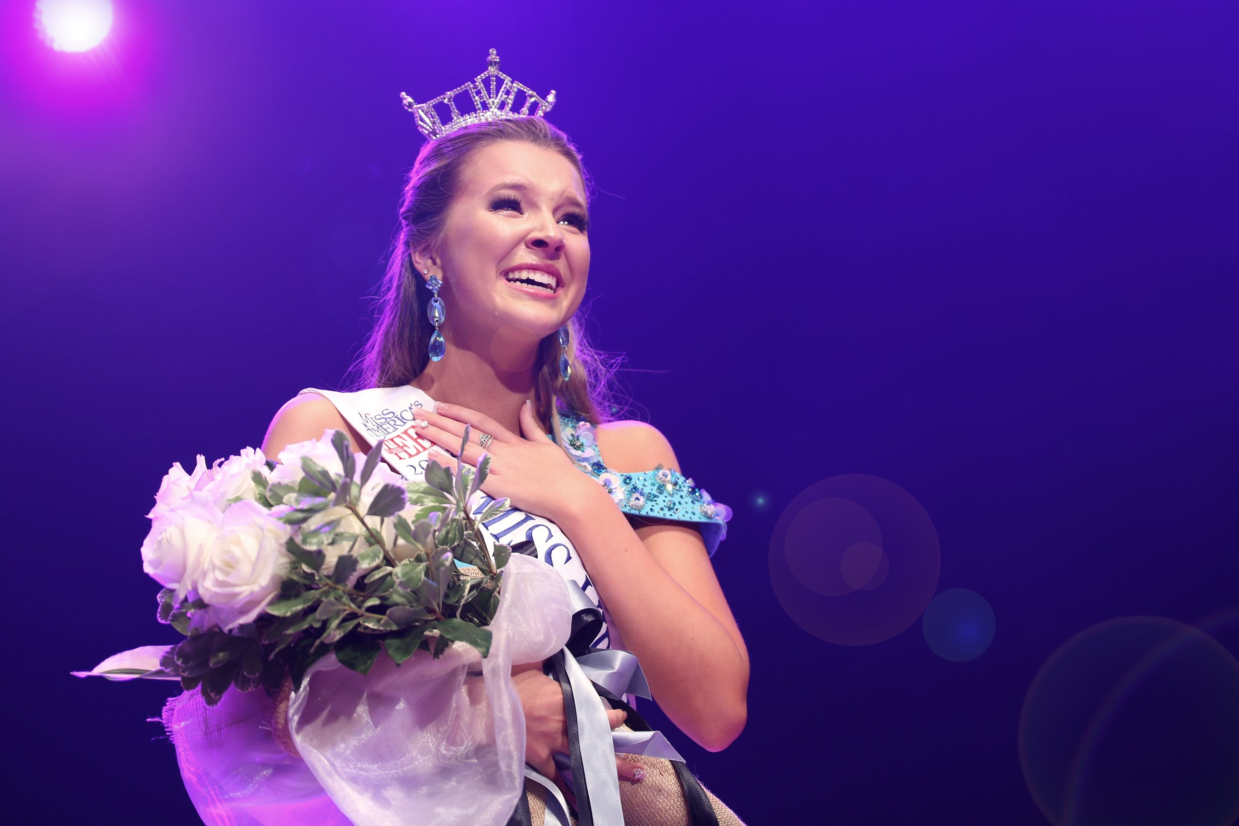 Miss America winner wearing a crown and sash, holding a bouquet of flowers, with a purple-lit stage background.