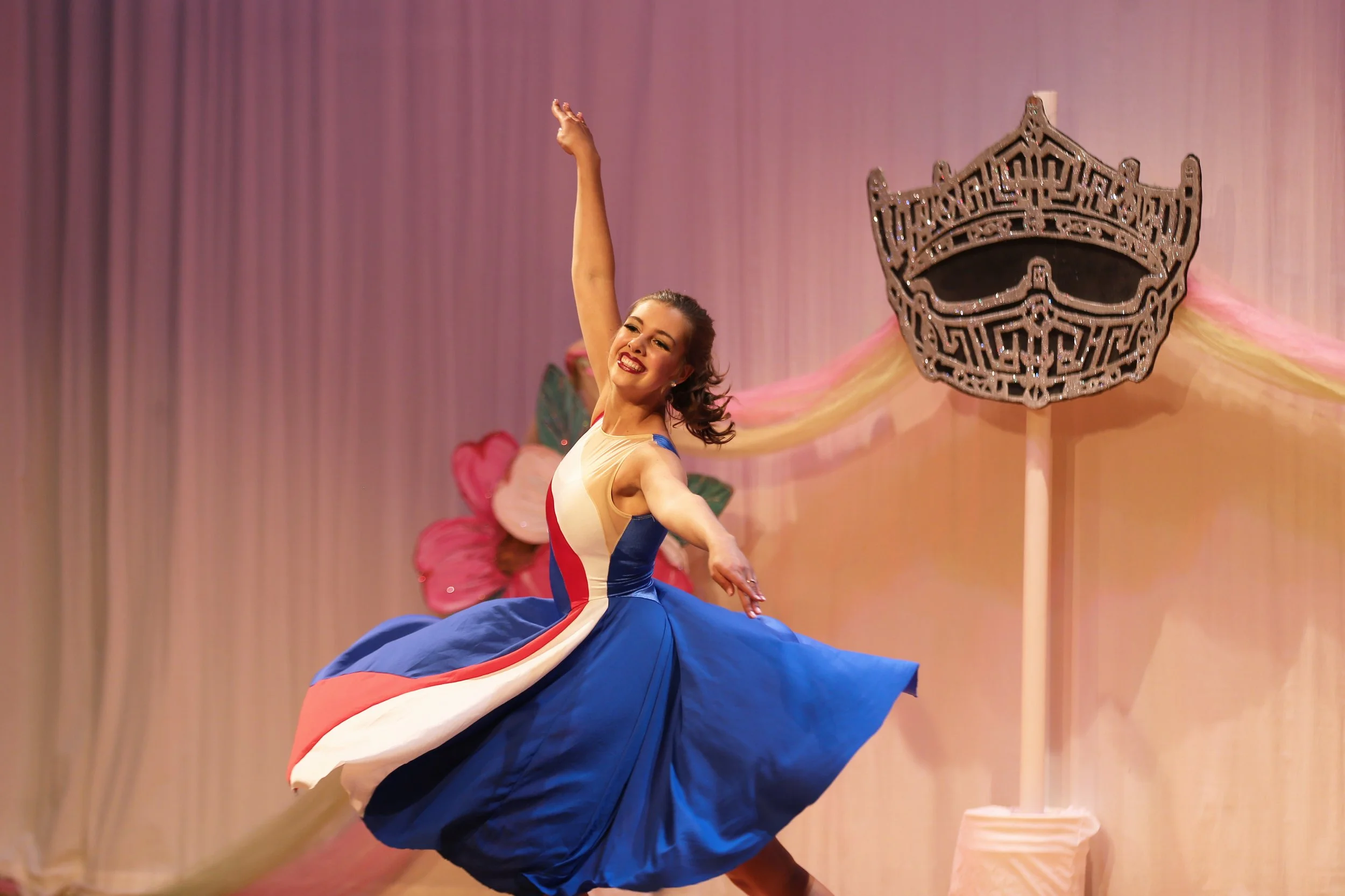 A woman in a colorful dress dancing happily on stage with large pink flowers and a crown-shaped sculpture in the background.