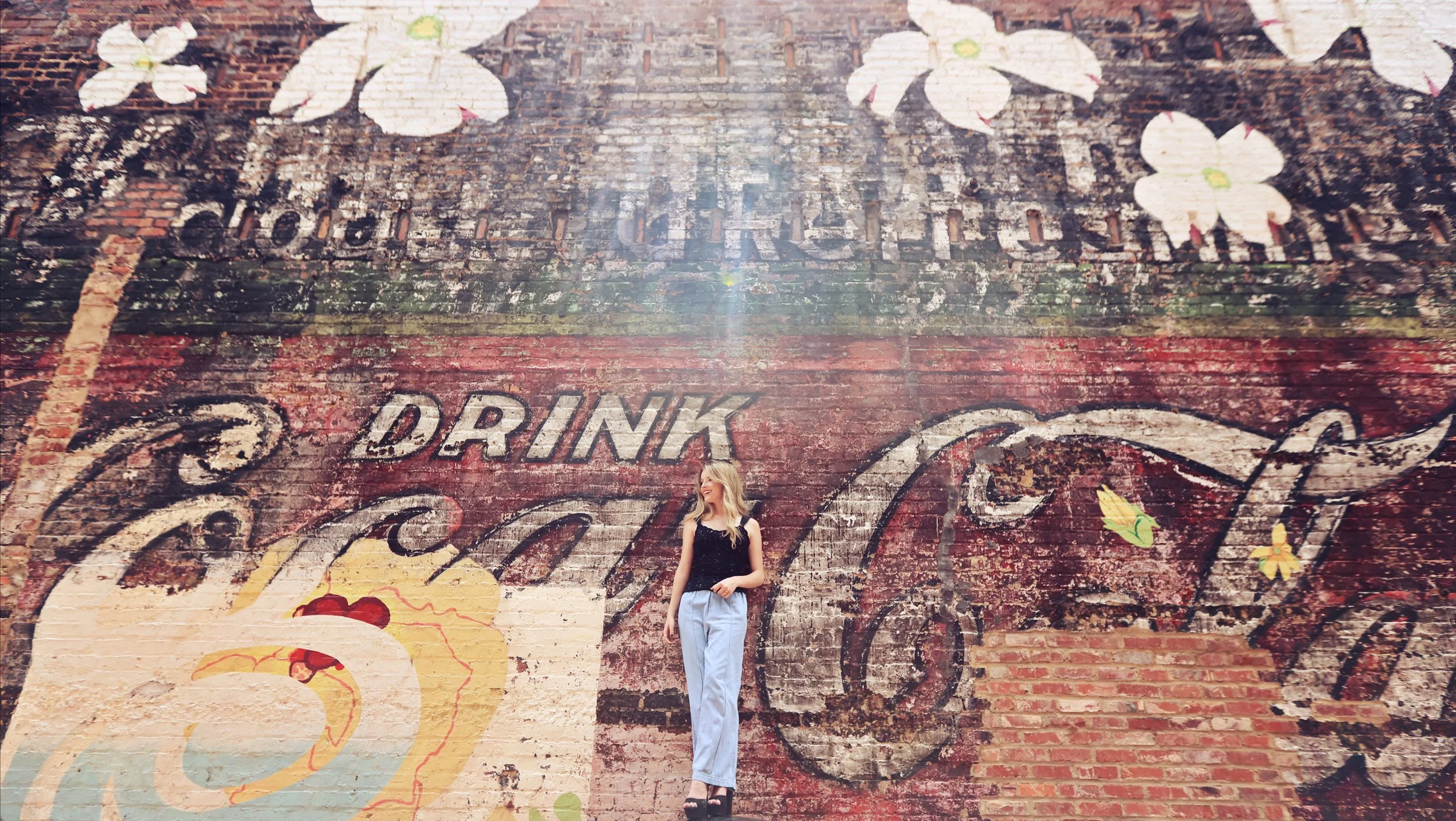 Woman standing in front of a large weathered brick wall with vintage Coca-Cola advertising and floral artwork, smiling and looking to her left.
