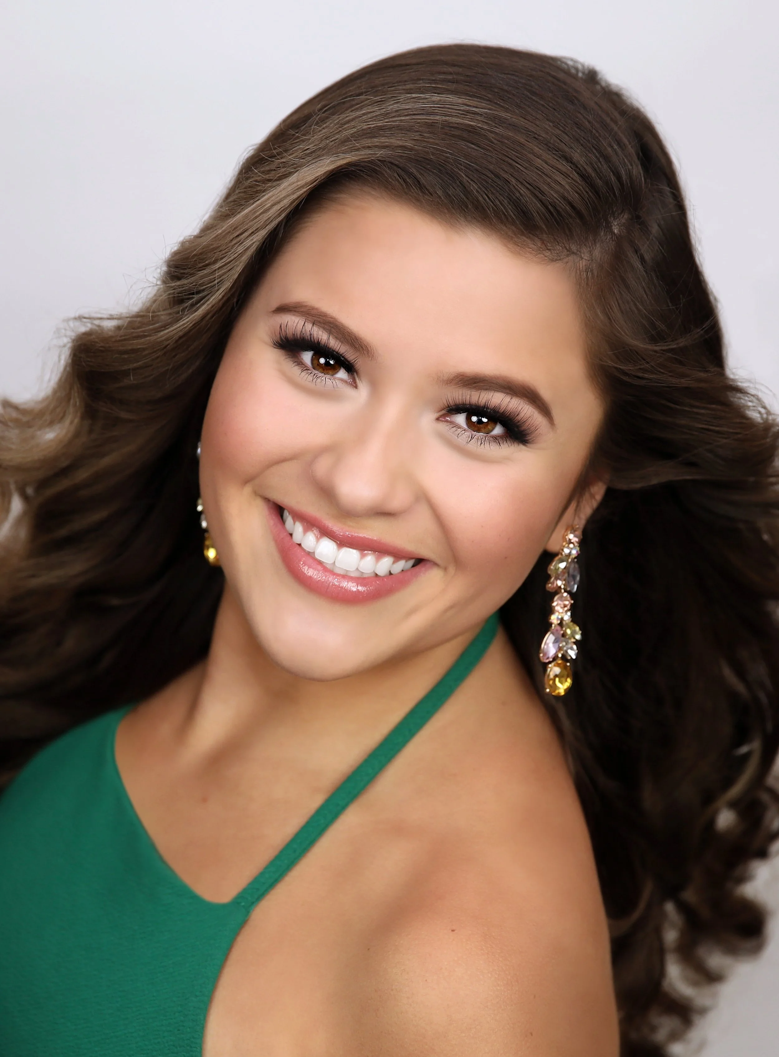 A smiling woman with brown, wavy hair wearing a green top and colorful, dangling earrings.
