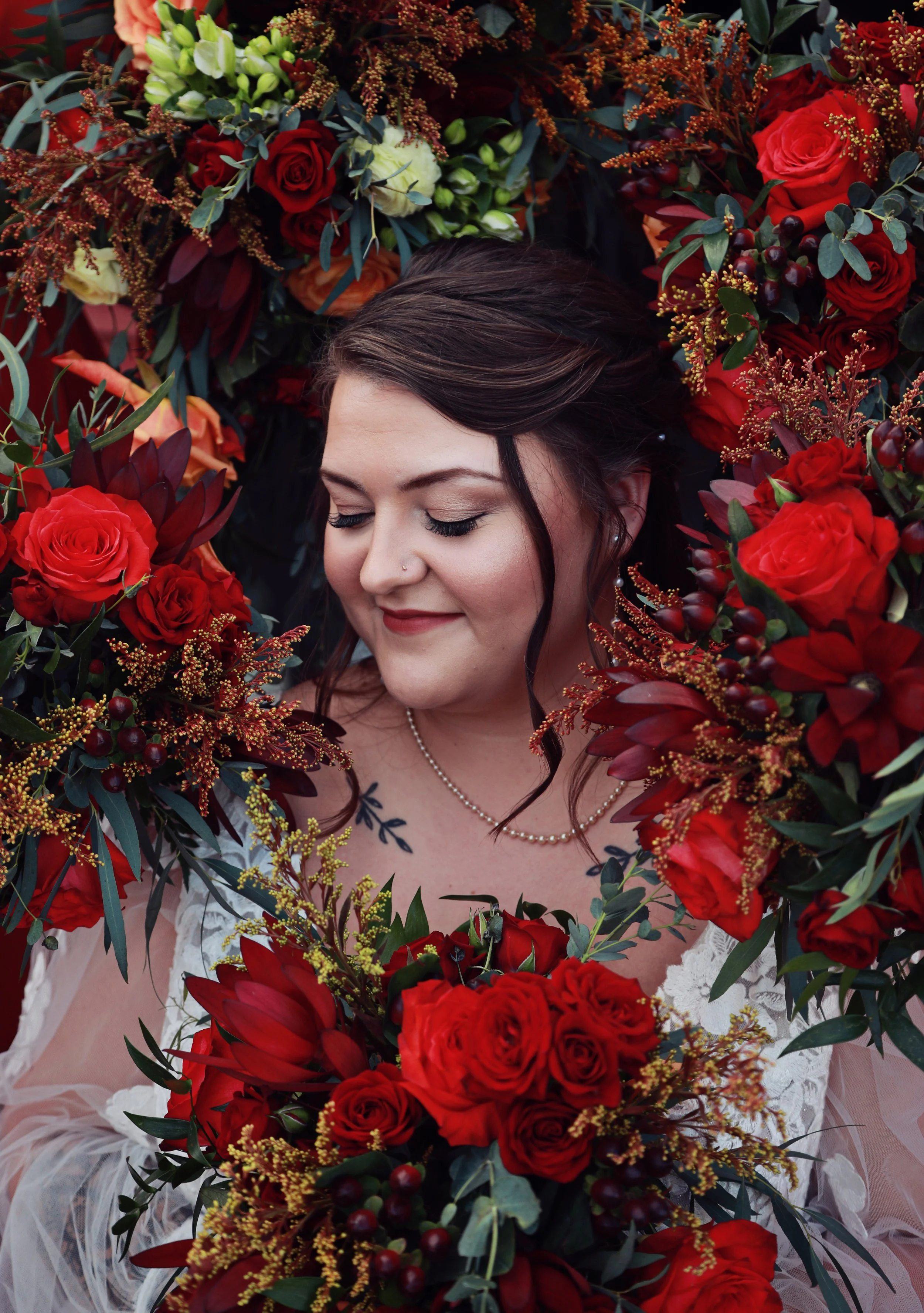 A woman with dark hair and makeup, wearing a white dress with lace and a pearl necklace, is surrounded by red roses, green leaves, and other colorful flowers, with a peaceful expression and eyes closed.
