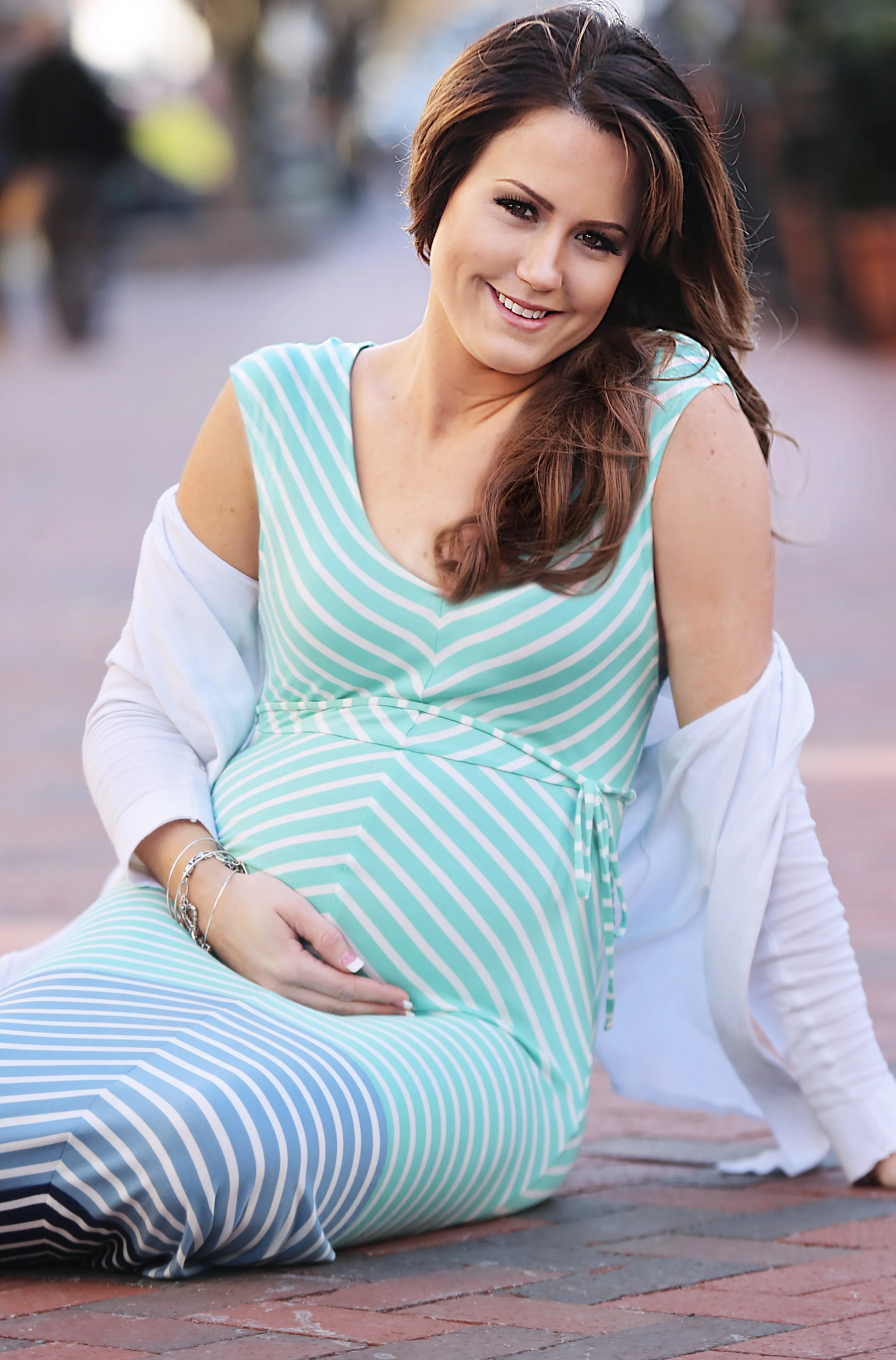 Pregnant woman with long brown hair sitting on the sidewalk, smiling, wearing a blue and white striped dress, and a white jacket draped over her shoulders.