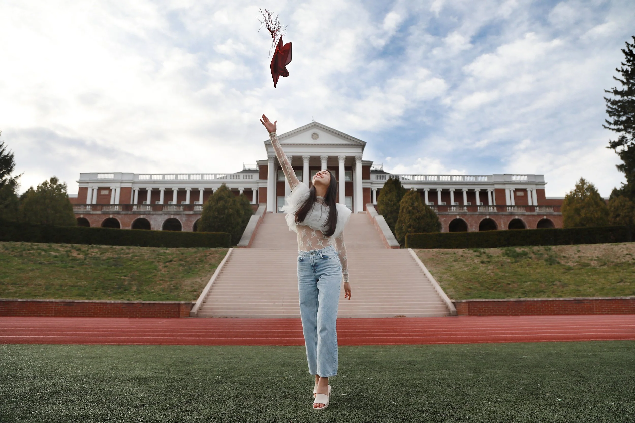 A young woman standing on a track at a university campus, throwing her graduation cap into the air, with a large white building featuring columns behind her and a partly cloudy sky overhead.