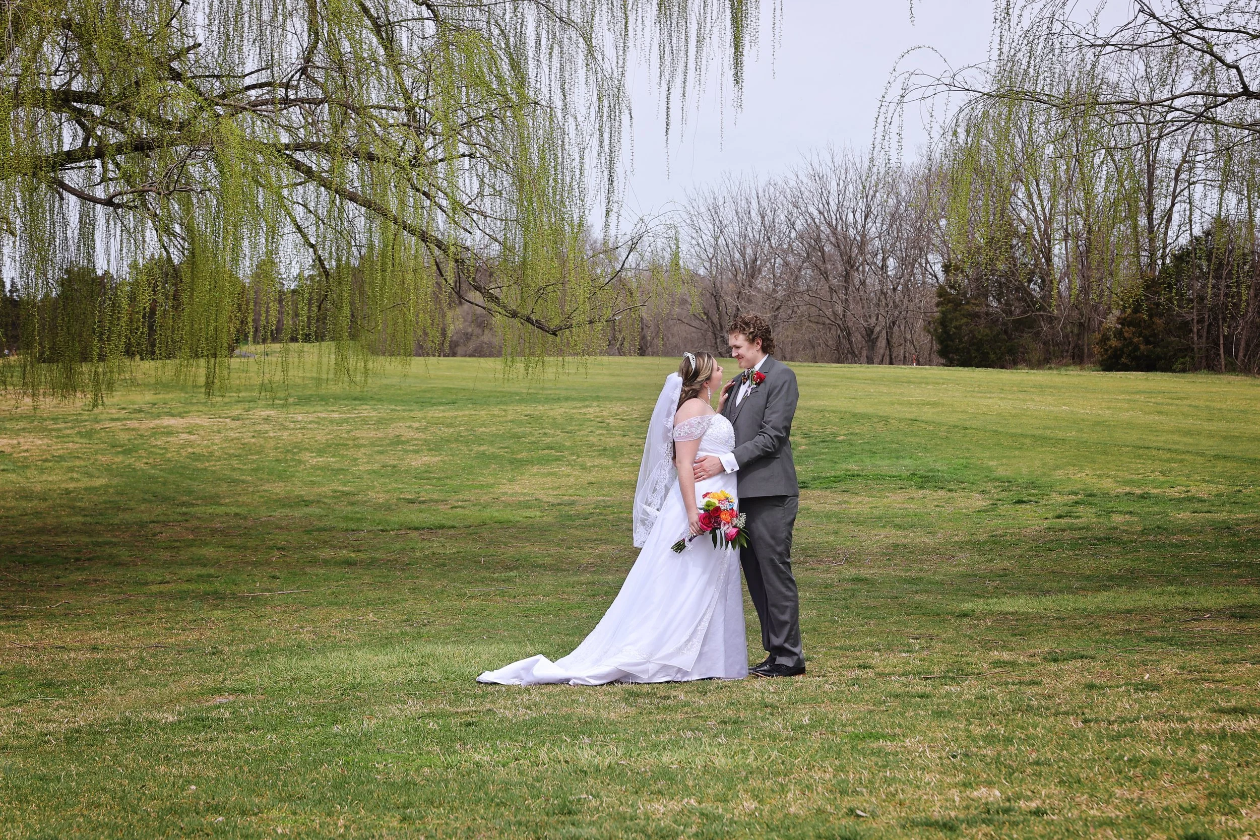A bride and groom stand together on a grassy field, under a tree with hanging branches, during their wedding photos. The bride wears a white wedding dress and holds a bouquet of red, pink, and yellow flowers. The groom wears a gray suit and a red boutonniere. The background features sparse trees with some greenery and budding leaves.