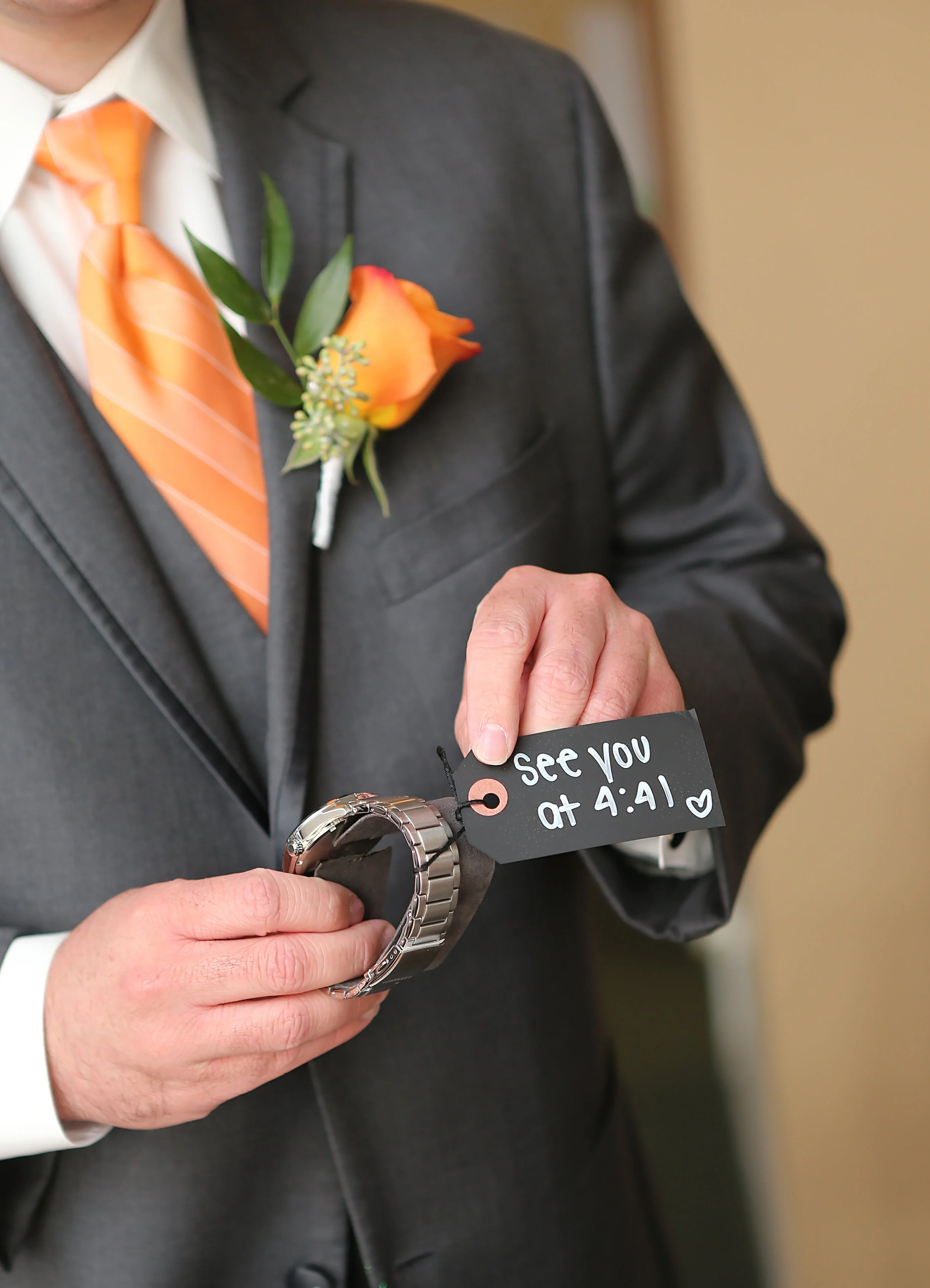 Person in a gray suit and orange striped tie holds a watch with a black tag that says "See you at 4:41" and a small heart. The person also has an orange flower boutonniere on their left lapel.