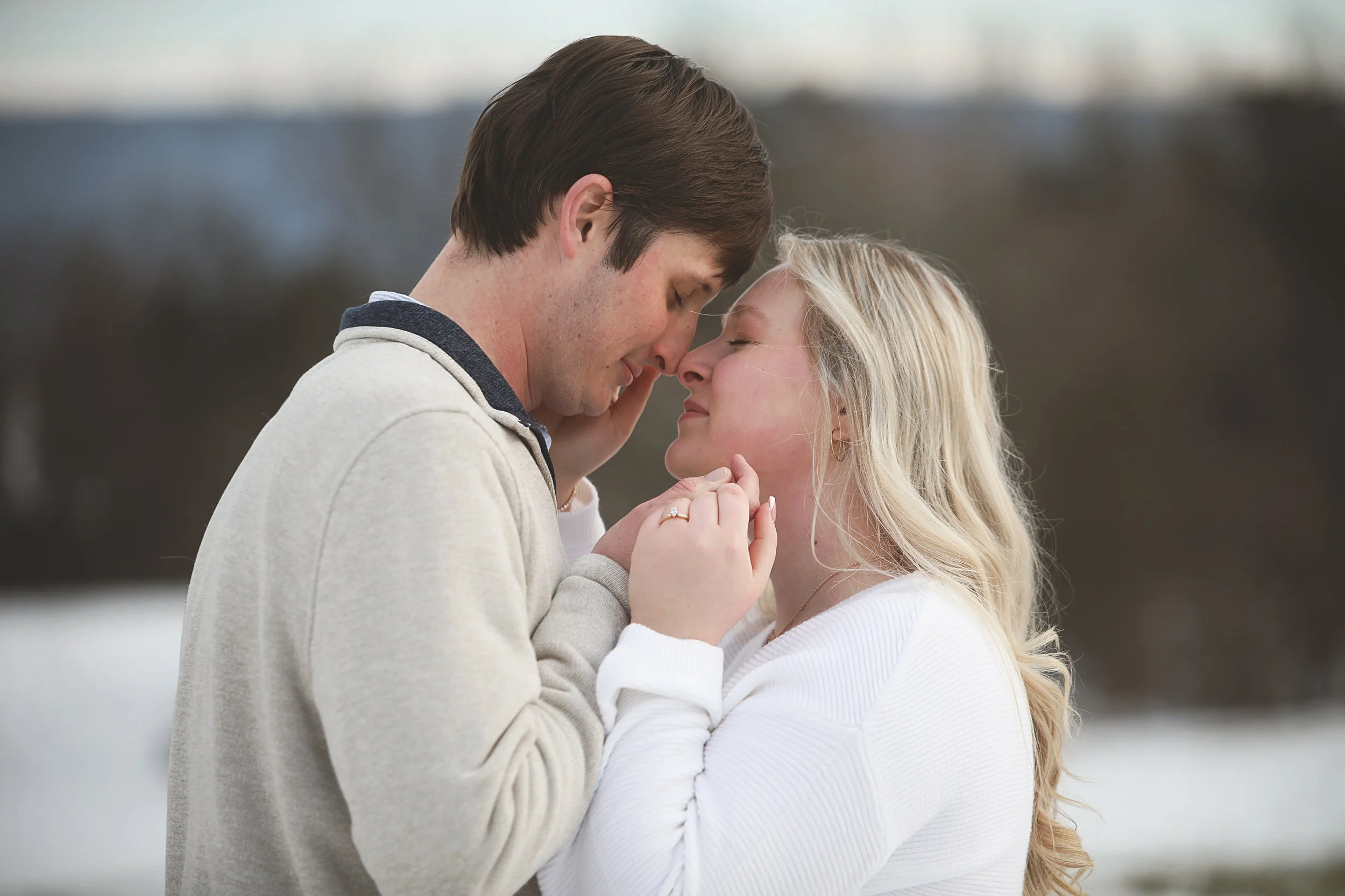A young couple sharing a close, intimate moment outdoors, with noses touching and eyes closed, her hands gently holding his face.