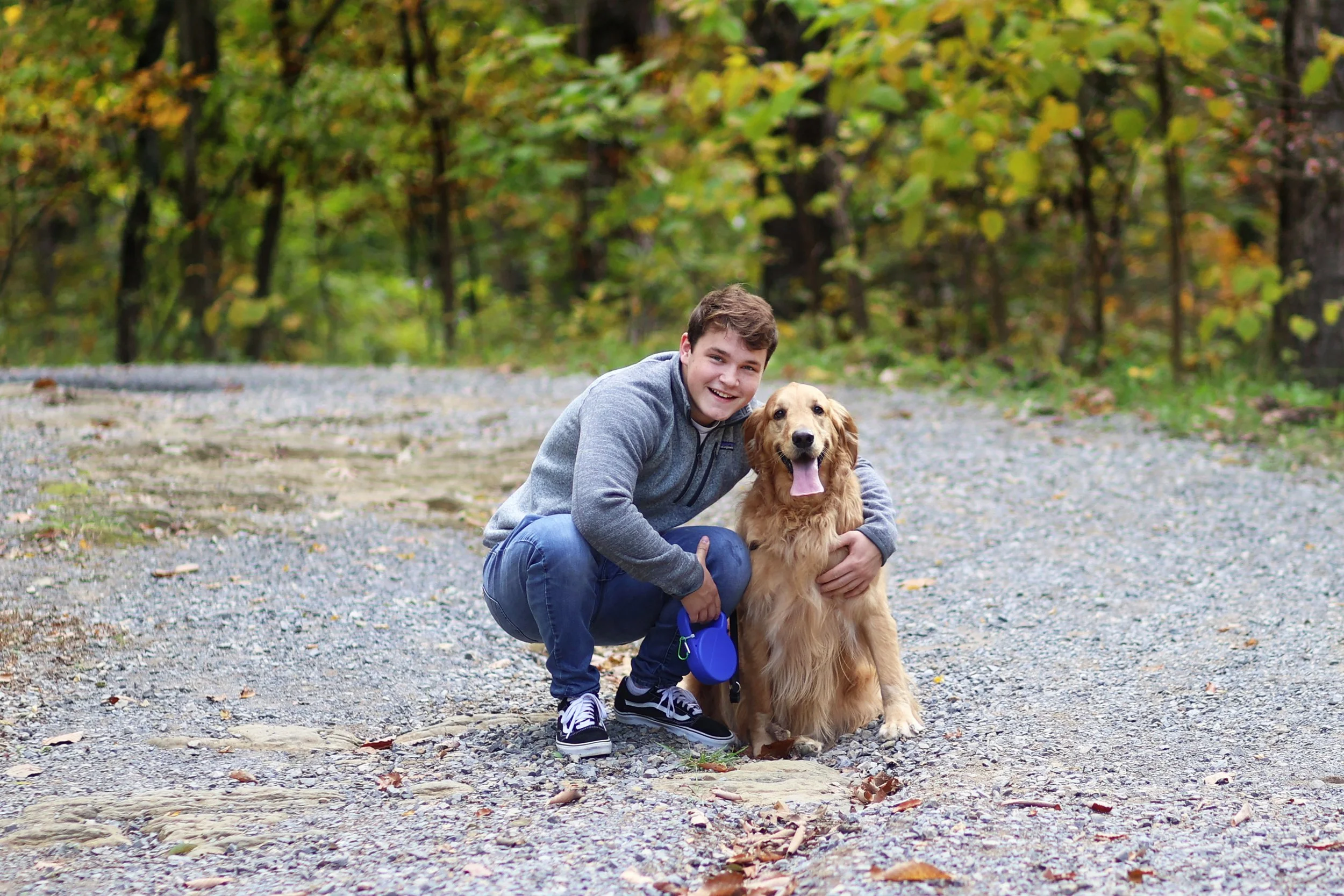 A young man crouching next to a golden retriever dog on a gravel path in a forest with green and yellow autumn leaves.