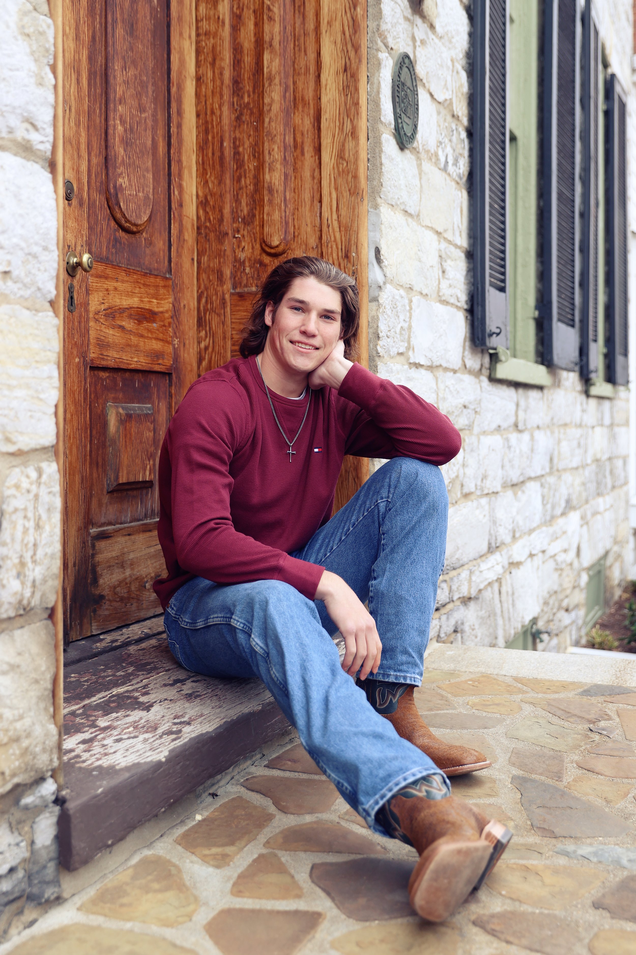 A young man with long brown hair, wearing a maroon long sleeve shirt, blue jeans, and cowboy boots, sitting on a stone step outside a house with a rustic wooden door and stone walls, smiling at the camera.
