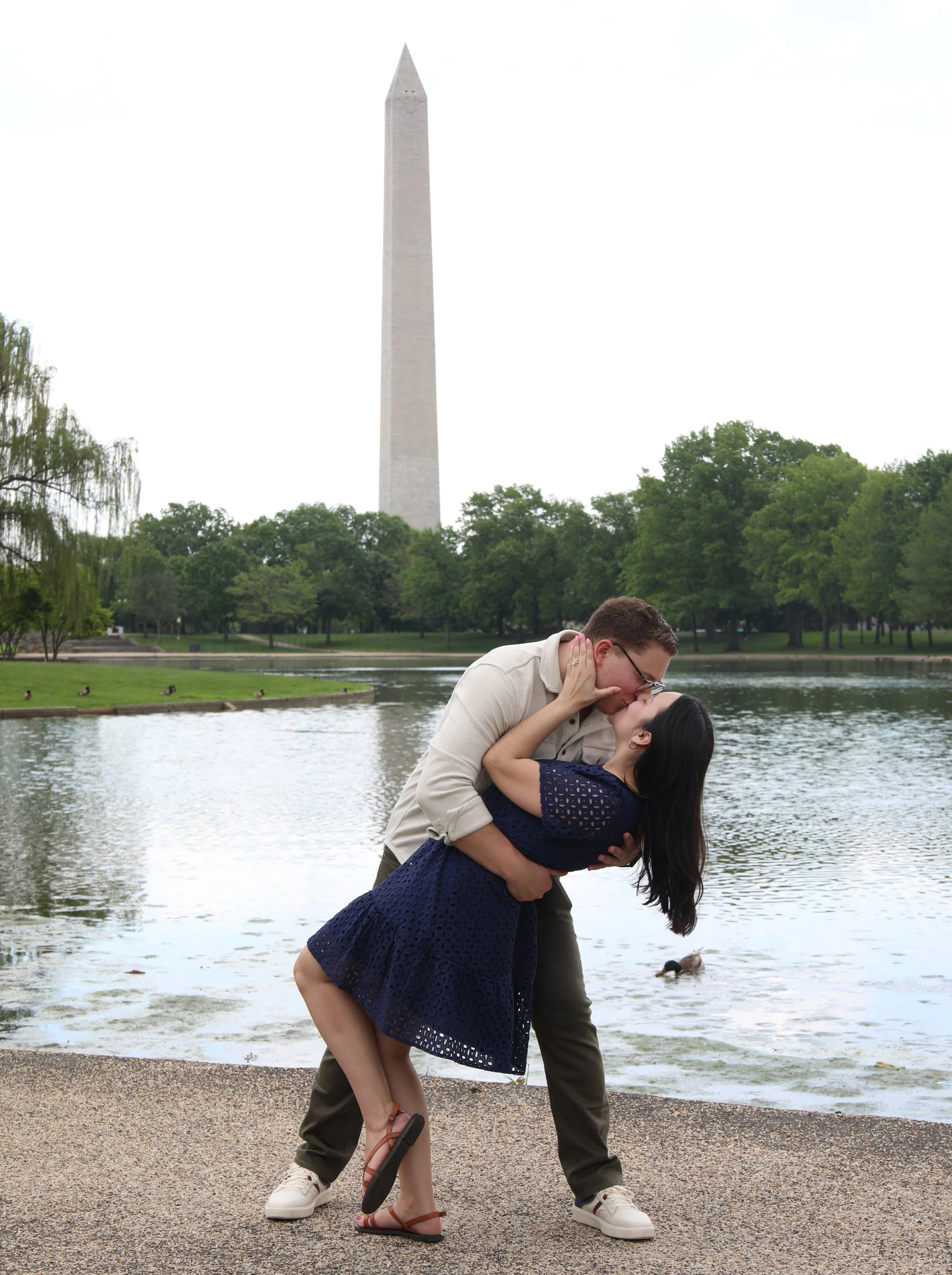 A couple kissing by a lake with the Washington Monument in the background, surrounded by trees.