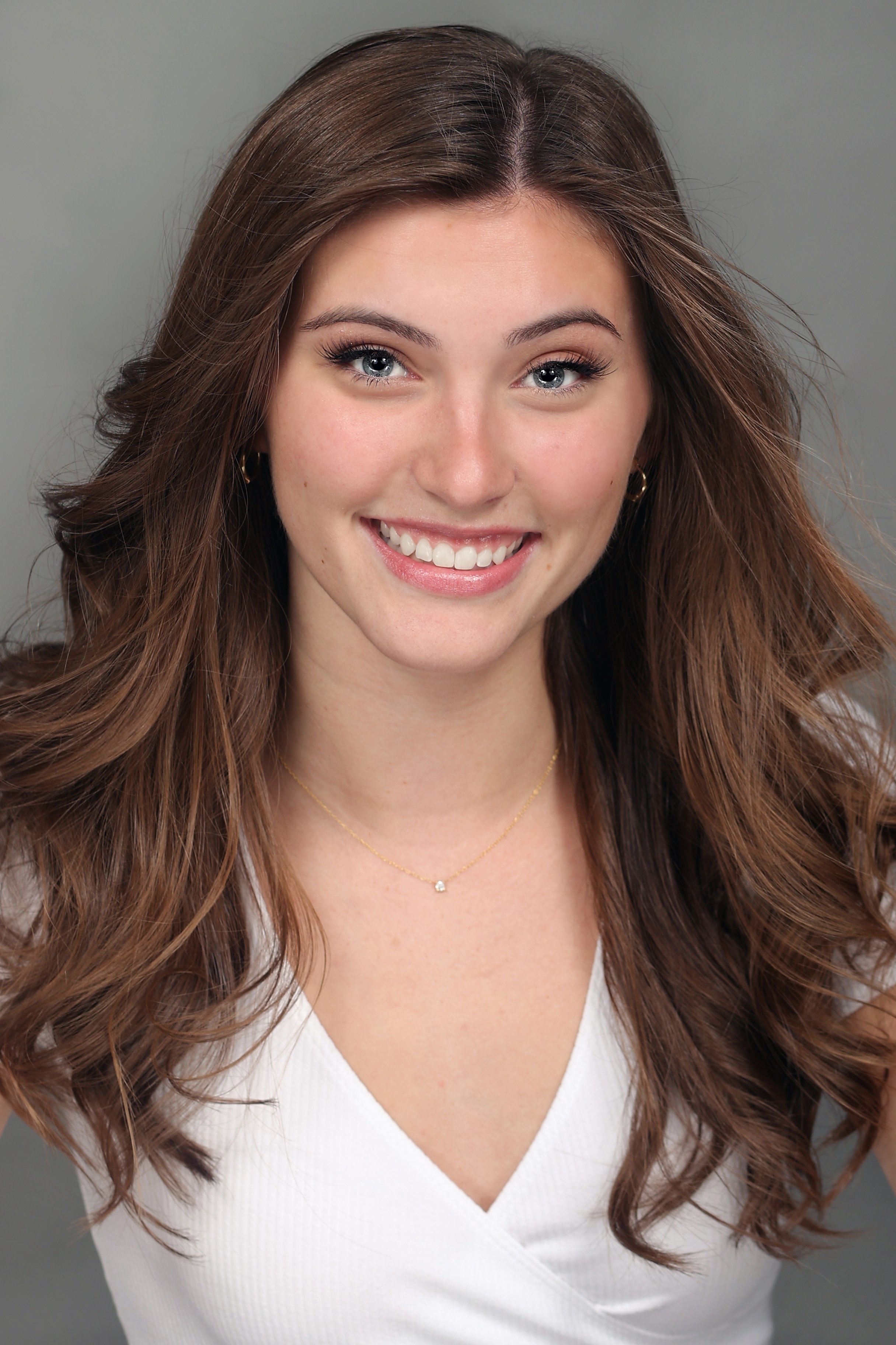 A young woman with long, wavy brown hair, blue eyes, and light skin, smiling at the camera, wearing a white top, gold earrings, and a delicate gold necklace with a small pendant, against a plain gray background.