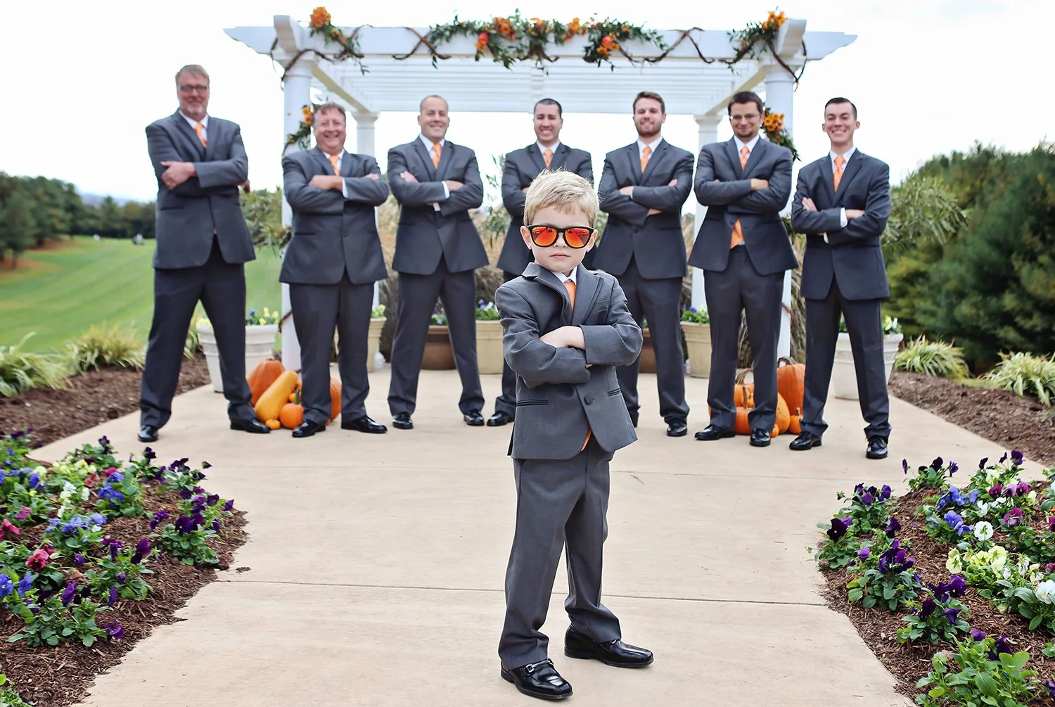 A young boy in sunglasses and a suit with arms crossed standing in front of a group of eight men dressed in suits and orange ties, at an outdoor wedding ceremony with autumn decorations and pumpkins.