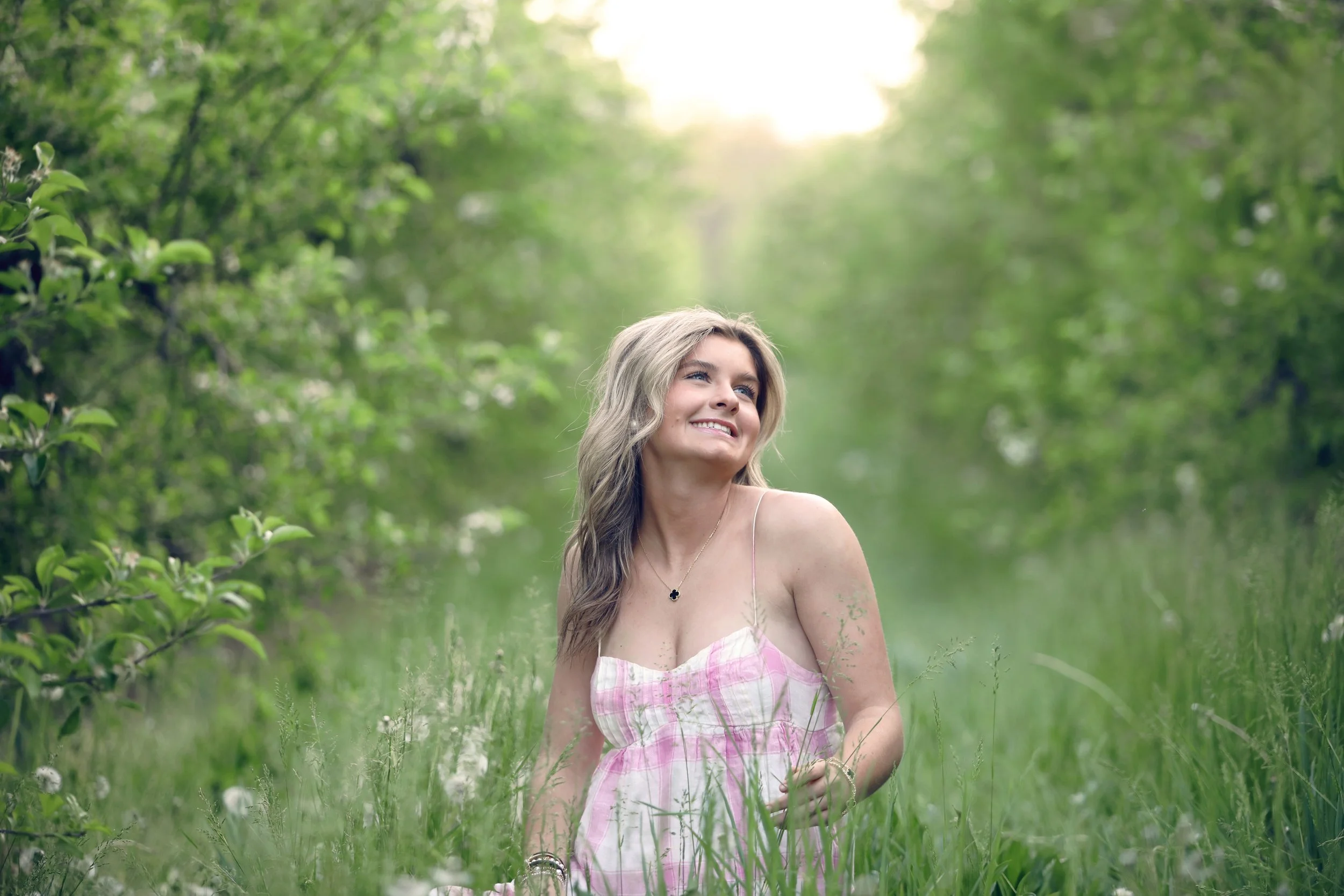 A woman with long blonde hair wearing a pink and white dress, smiling and sitting in a lush green outdoor setting with trees and grass.