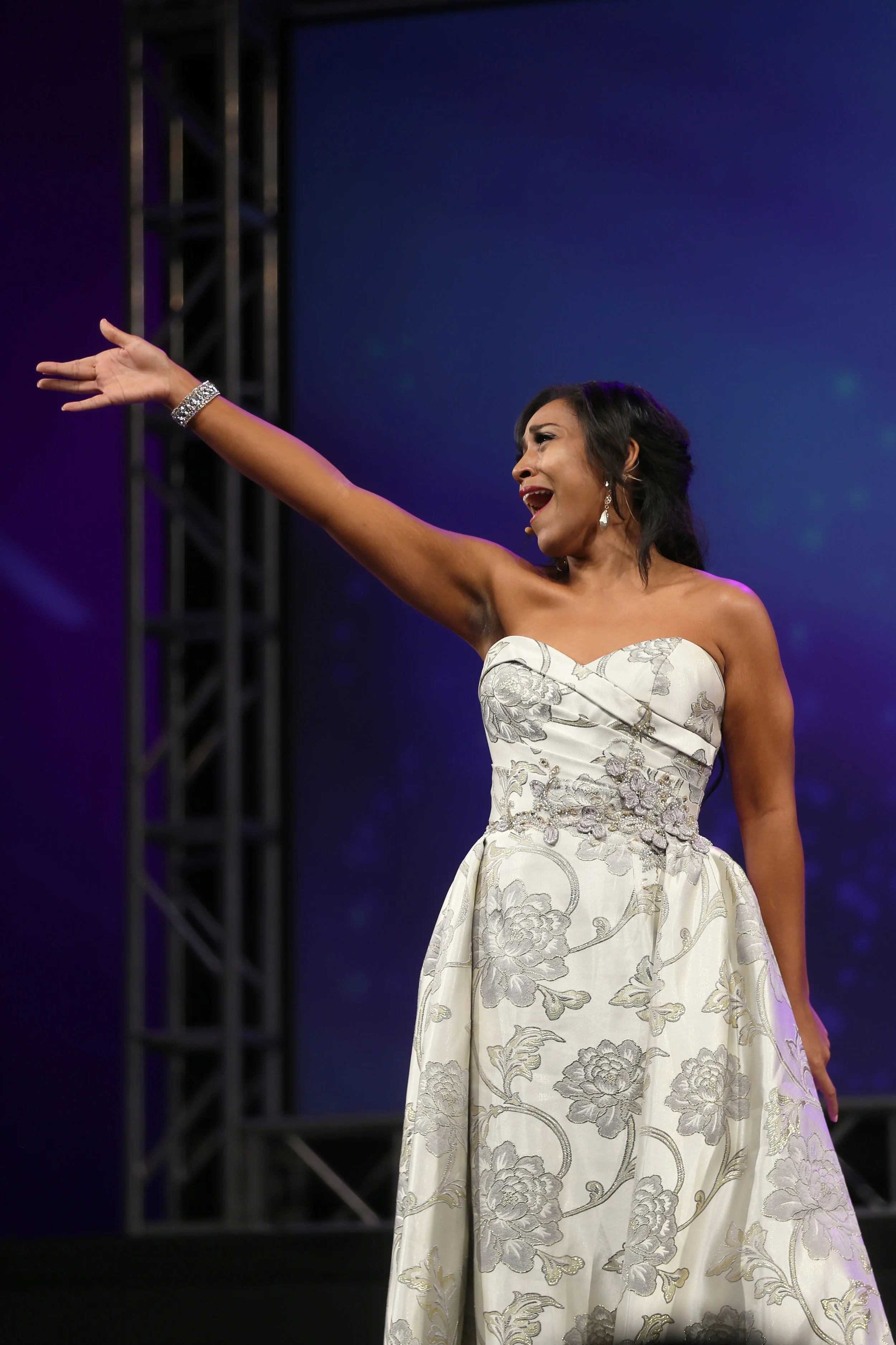 A woman in a strapless, floral-patterned gown is performing or speaking on stage, with her right arm extended outward and her mouth open.
