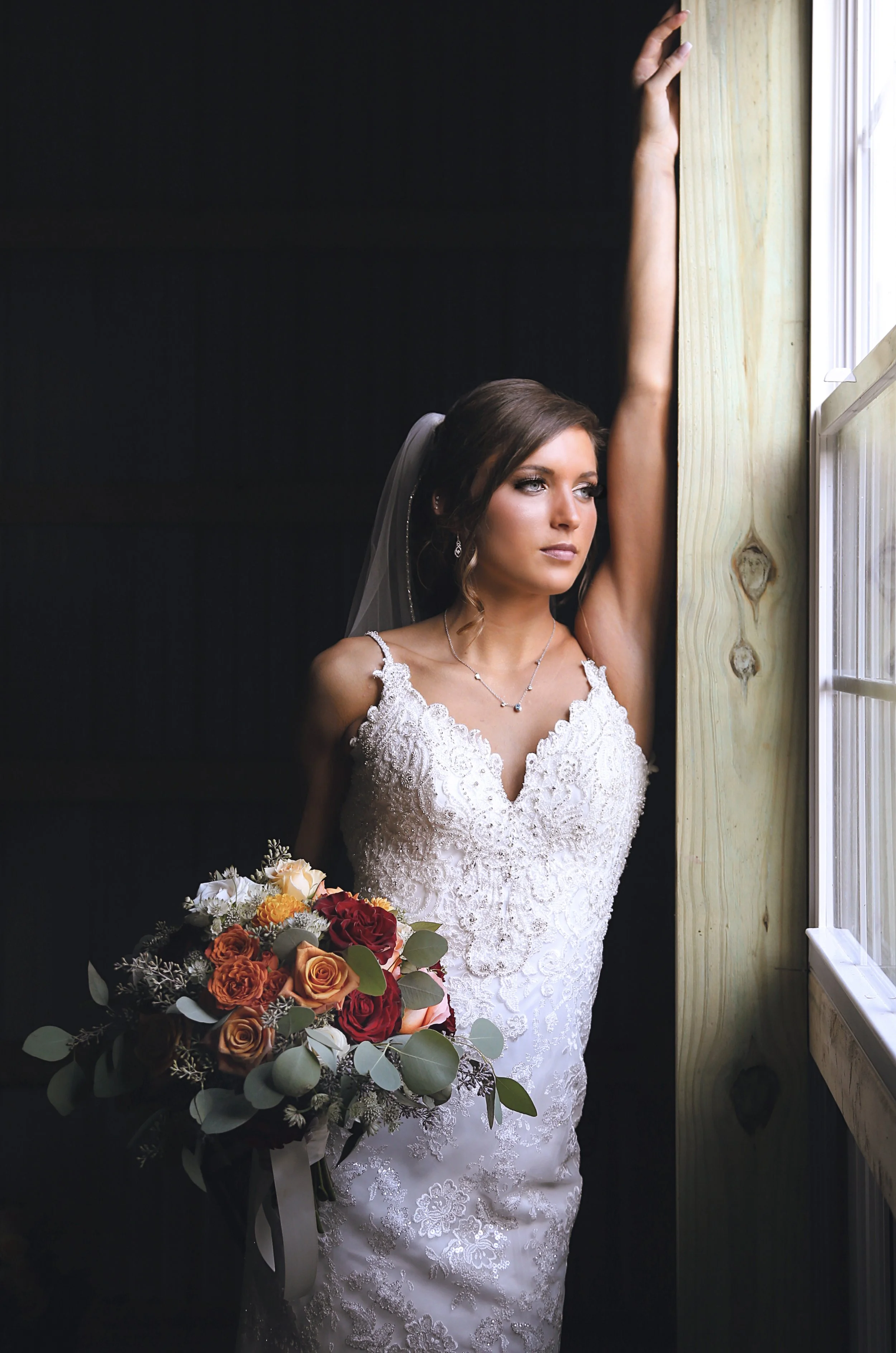 A bride in a white lace wedding dress holding a bouquet of roses, standing by a window with her arm raised, looking contemplative.