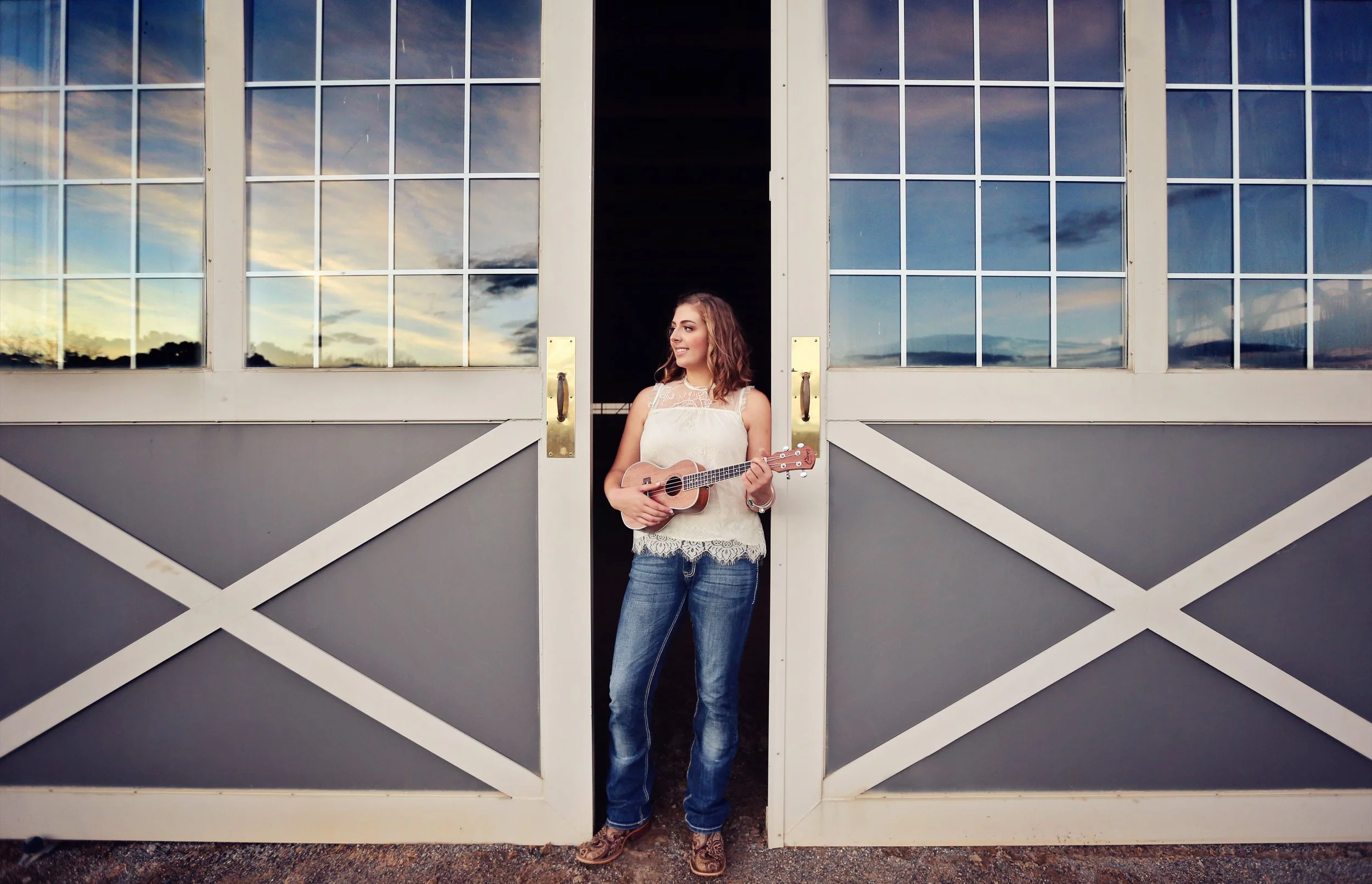 A young woman standing in a barn doorway, holding a ukulele, with sunset sky reflected in the glass windows behind her.