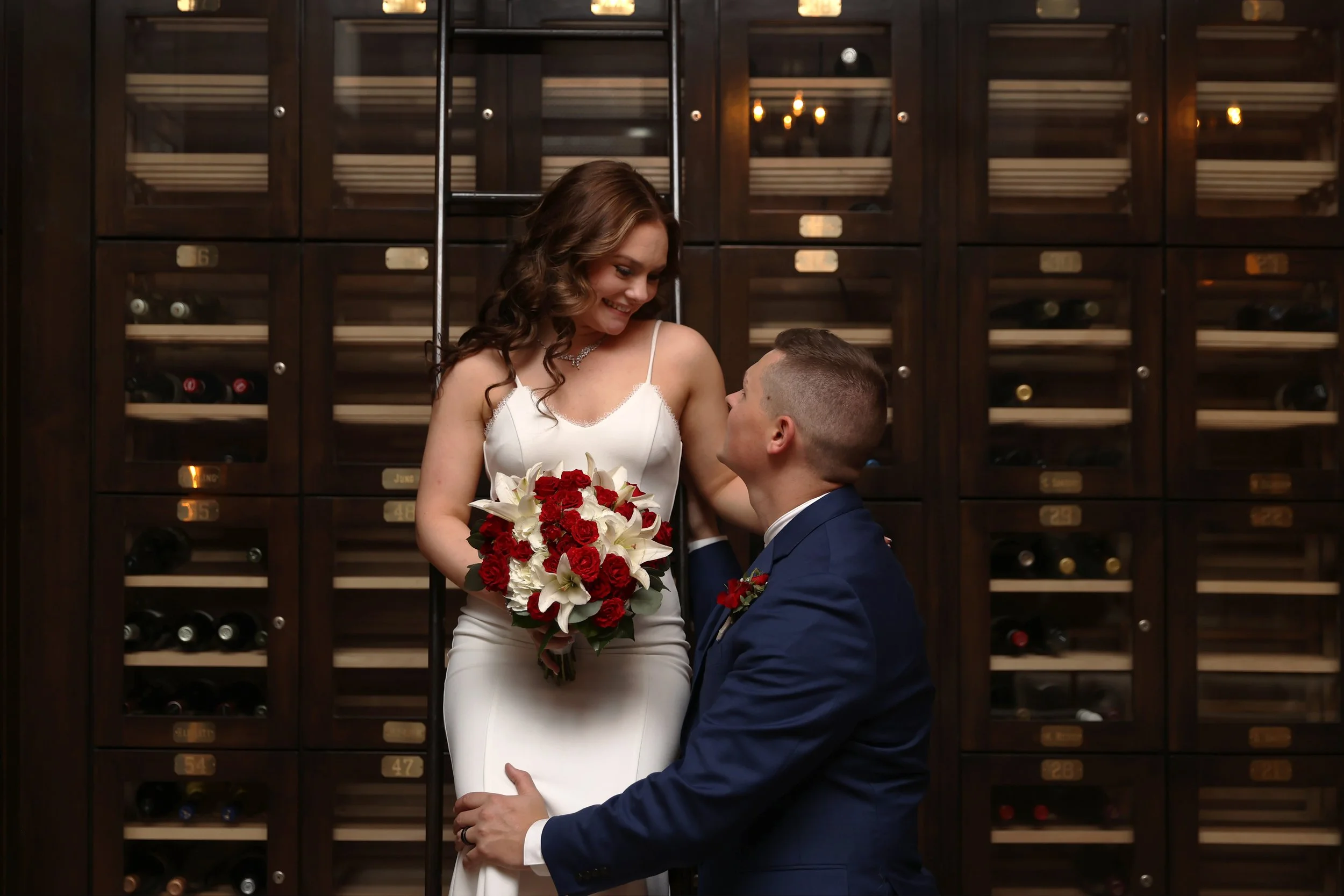 A bride and groom on their wedding day, standing in front of a wine cabinet. The bride wears a white dress and holds a bouquet of white lilies and red roses, while the groom wears a navy blue suit with a red boutonniere. They look at each other affectionately.
