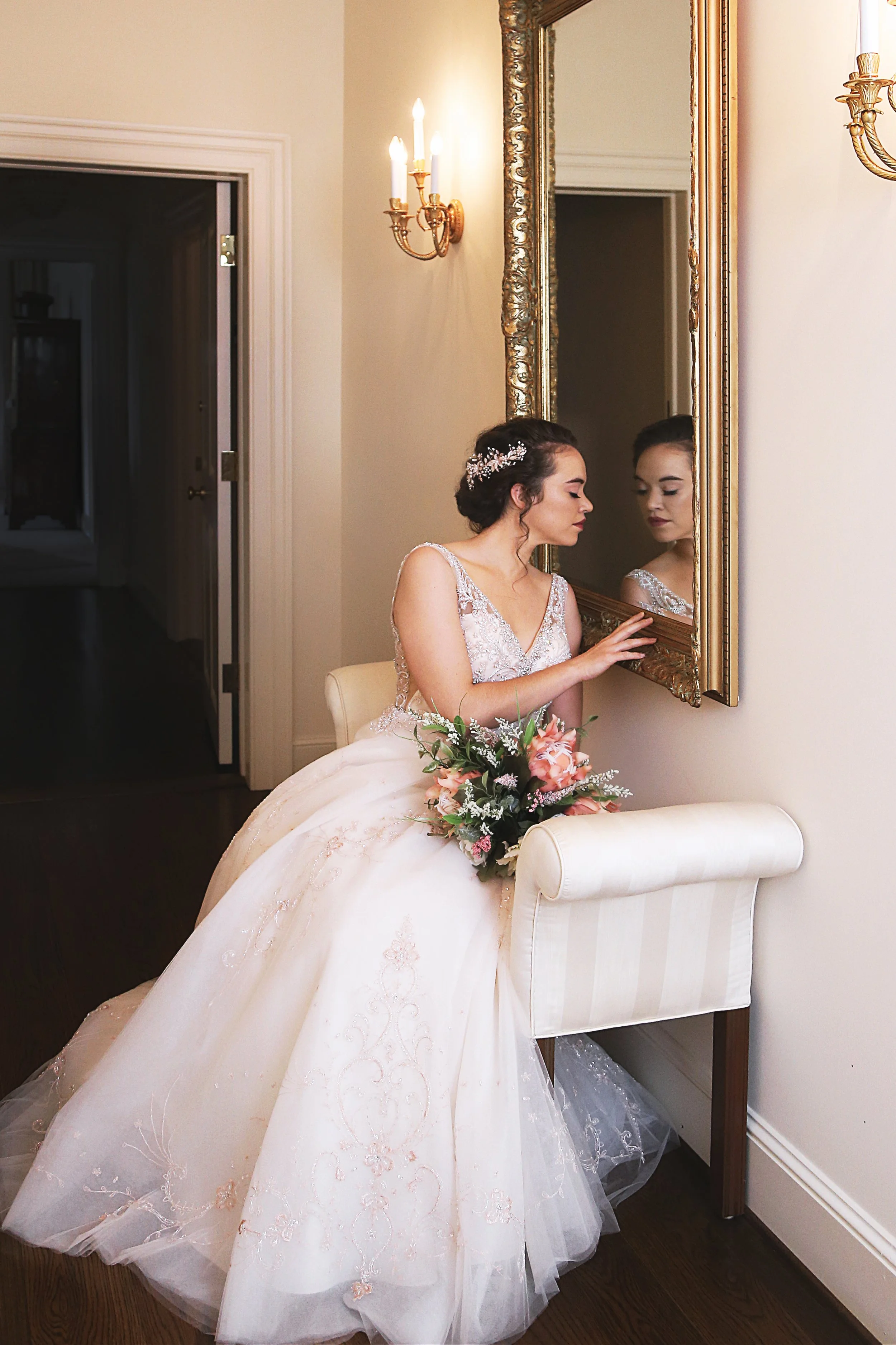 A bride in a wedding dress touching a large ornate mirror, holding a bouquet of pink flowers, in a well-lit room.