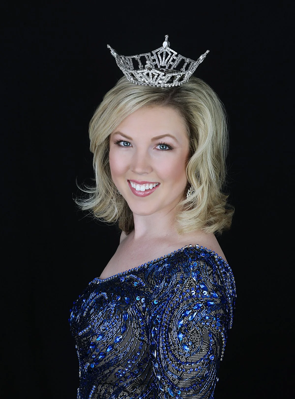 A woman with blonde hair and blue eyes wearing a blue sequined dress, a silver tiara, and earrings, posing against a black background.