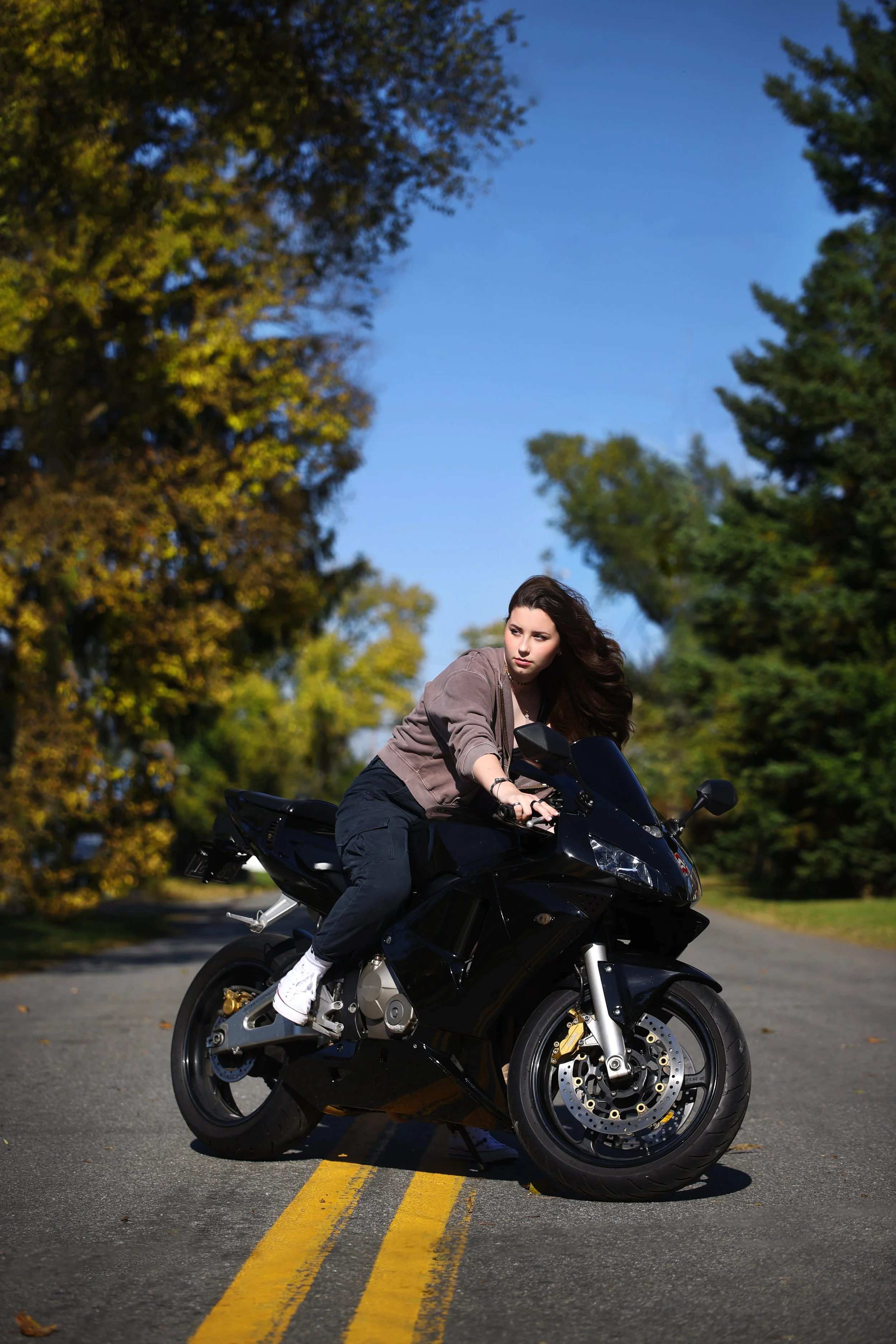 A young woman riding a black motorcycle on a road surrounded by trees during daytime.