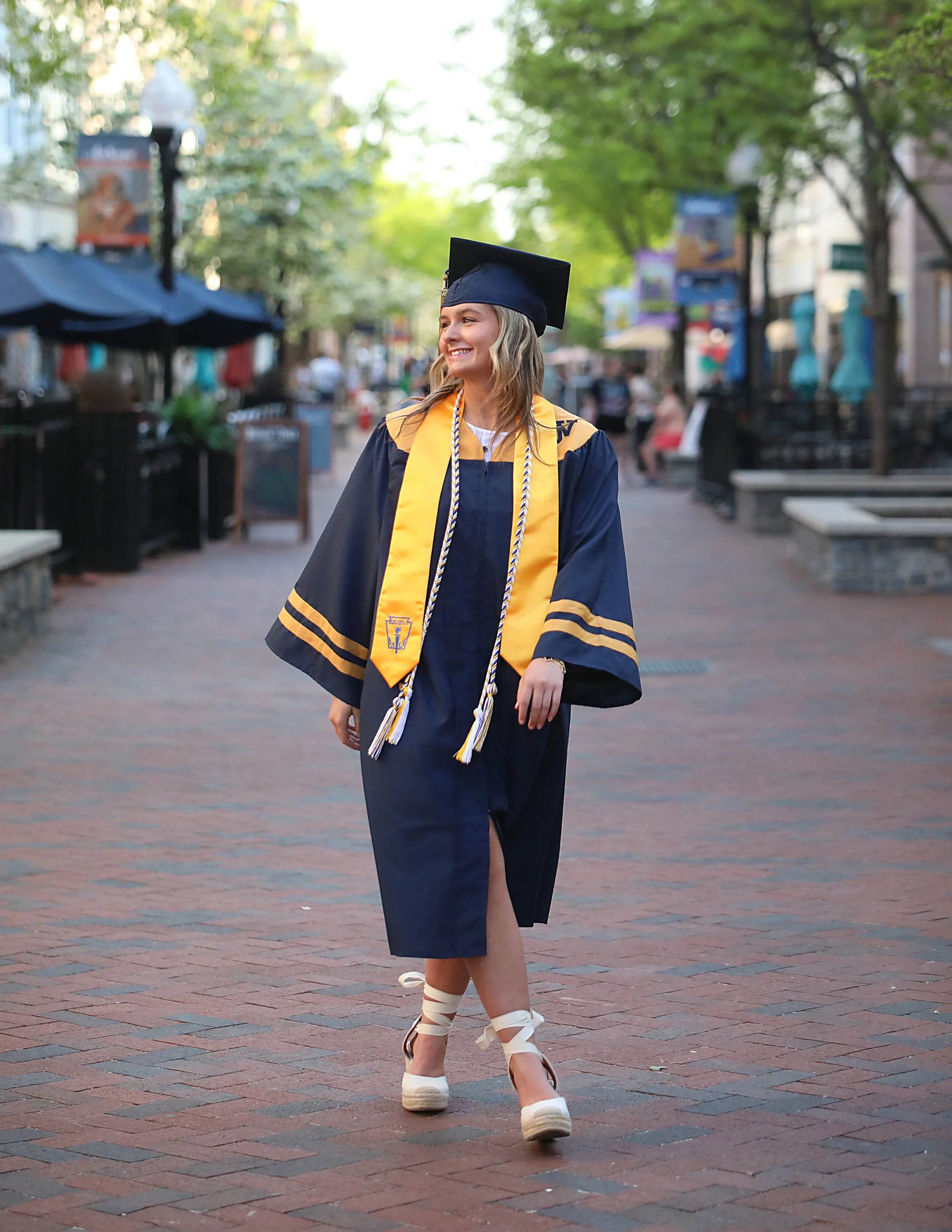 A young woman in a graduation cap and gown walking on a brick-paved street, smiling, with trees and outdoor seating in the background.