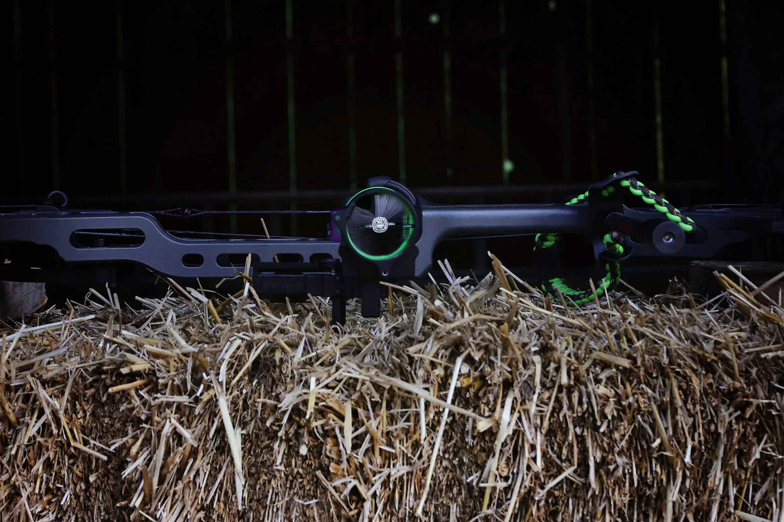 A black and green crossbow resting on a hay bale in a dark setting.
