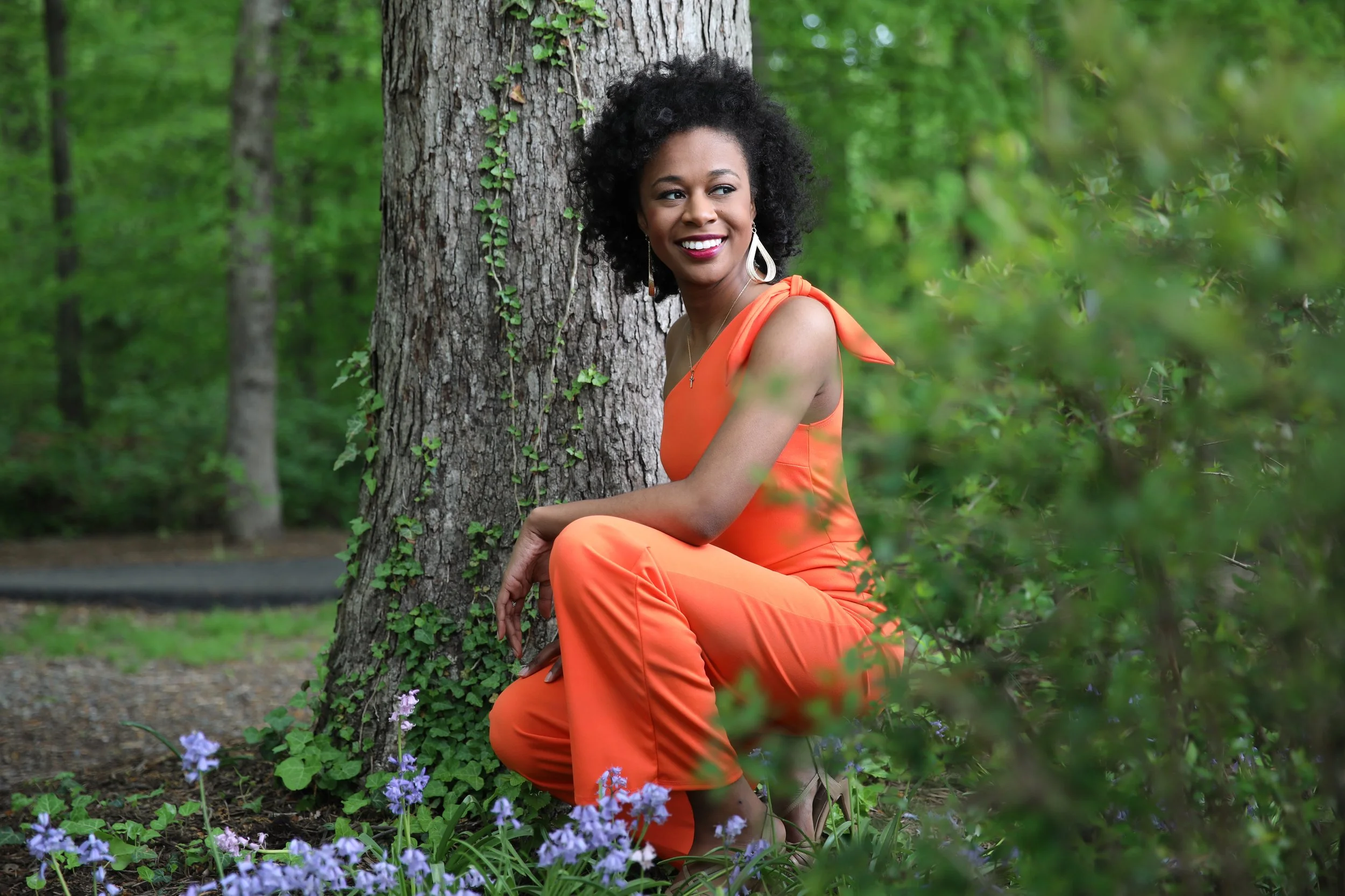 A woman in an orange dress kneels beside a tree in a green forest, smiling and looking to the side.