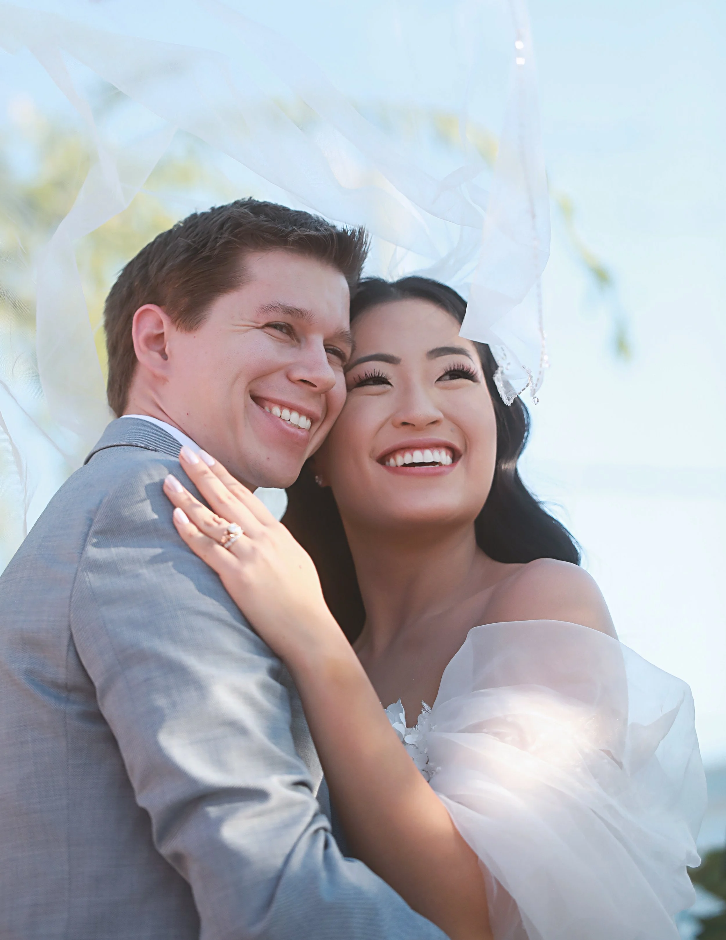 A happy couple embracing outdoors, smiling, with a veil and wedding attire, under a white canopy or umbrella.