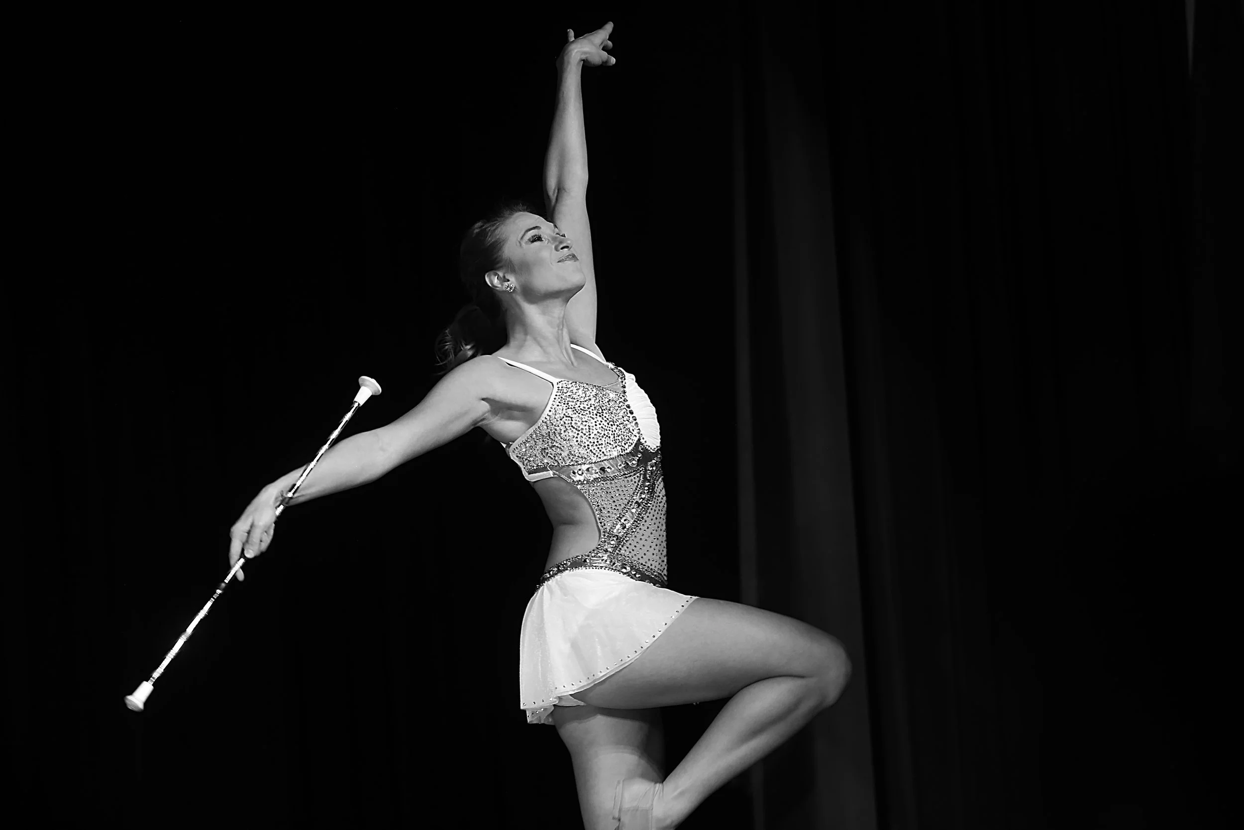 A female rhythmic gymnast performing a routine on stage, balancing on one leg with her other leg lifted, holding a club in one hand, wearing a glittering leotard and skirt, in front of dark curtains.