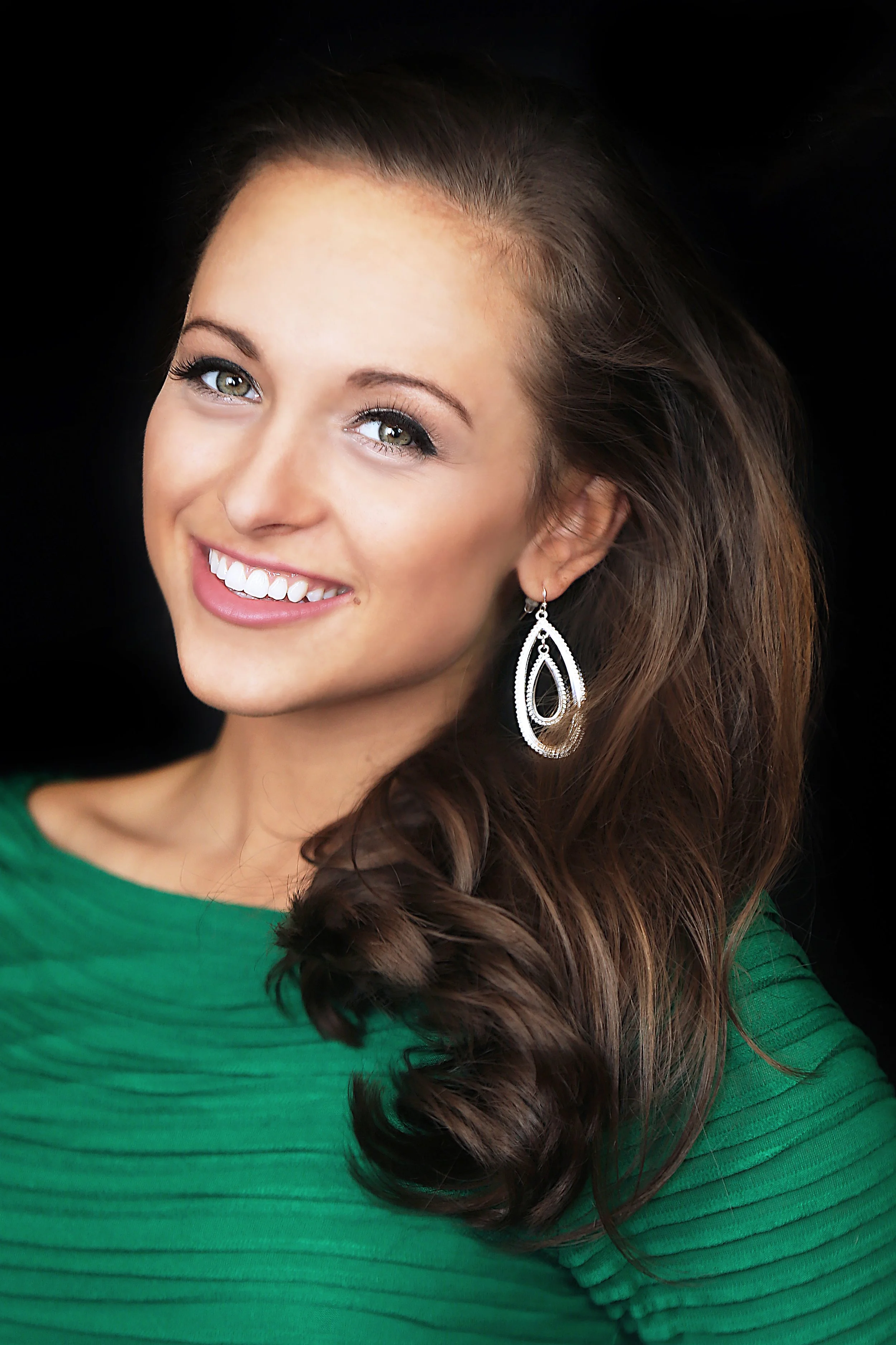 Close-up of a smiling woman with wavy brown hair, wearing a green top and large silver teardrop-shaped earrings against a black background.